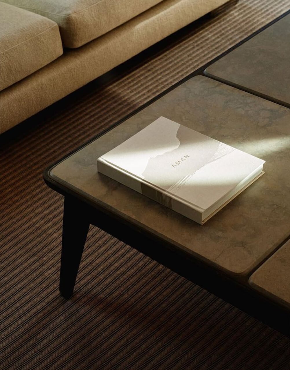 Aman New York, North America — Minimalist coffee table with cream-bound book beside camel sofa and textured area rug in luxury hotel lobby.