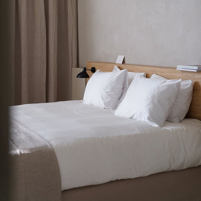 Hotel âme, Europe — minimalist bedroom with natural light, wooden headboard, crisp white linens, layered pillows, black reading lamp, neutral linen curtains, and serene hospitality design.