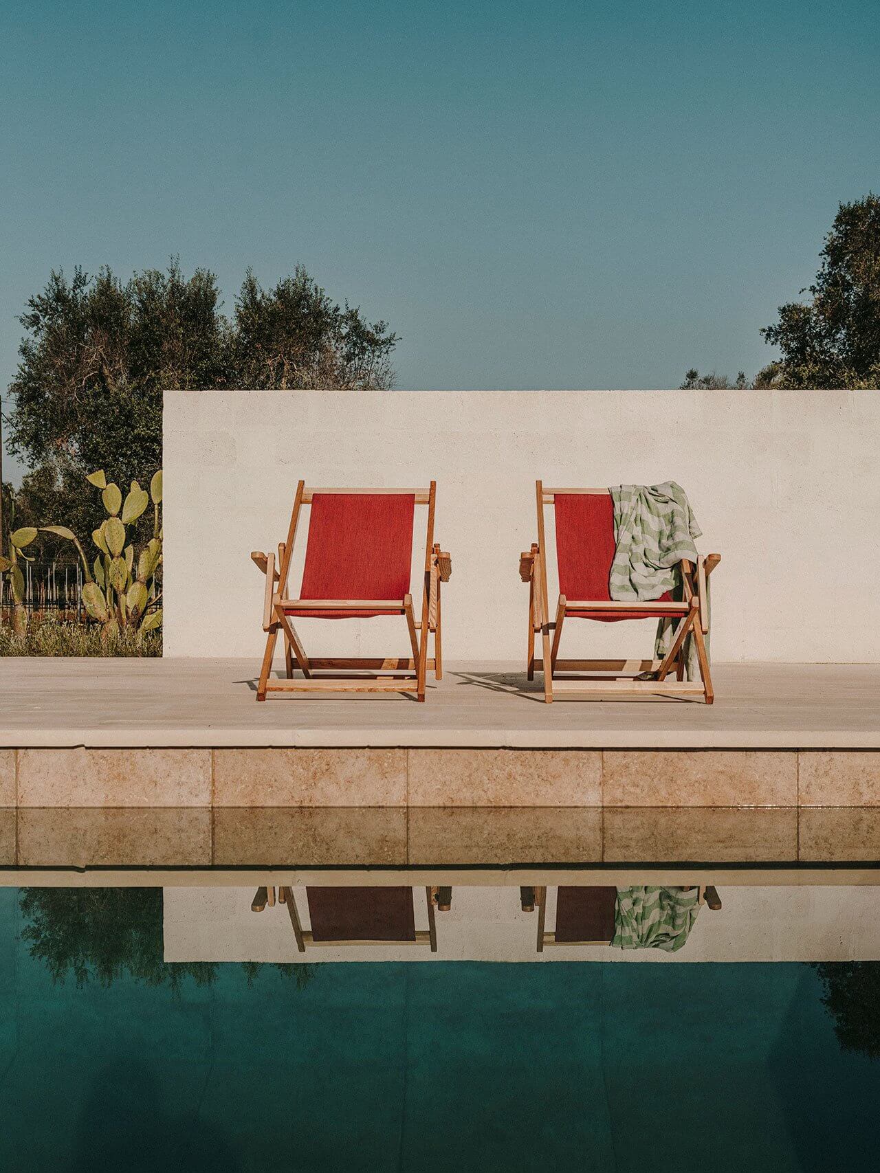 Borgo Gallana, Europe — Two wooden deck chairs with red fabric on stone terrace overlooking turquoise pool, white wall and cypress trees beyond.