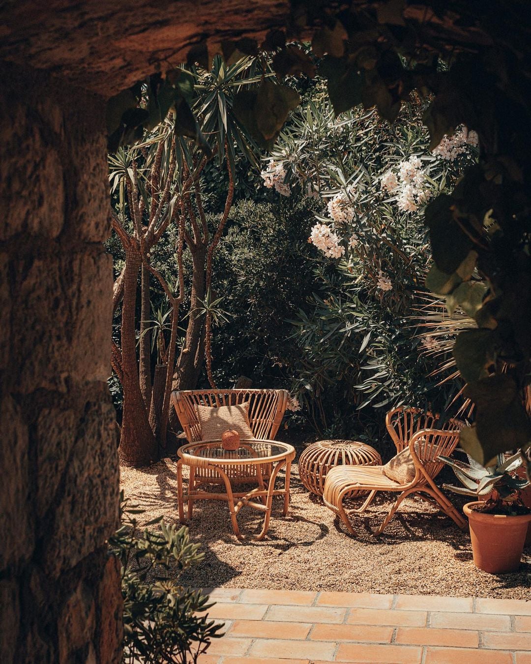 Epi 1959, Europe — Woven rattan chairs and table in candlelit courtyard garden with flowering plants and terracotta walls.