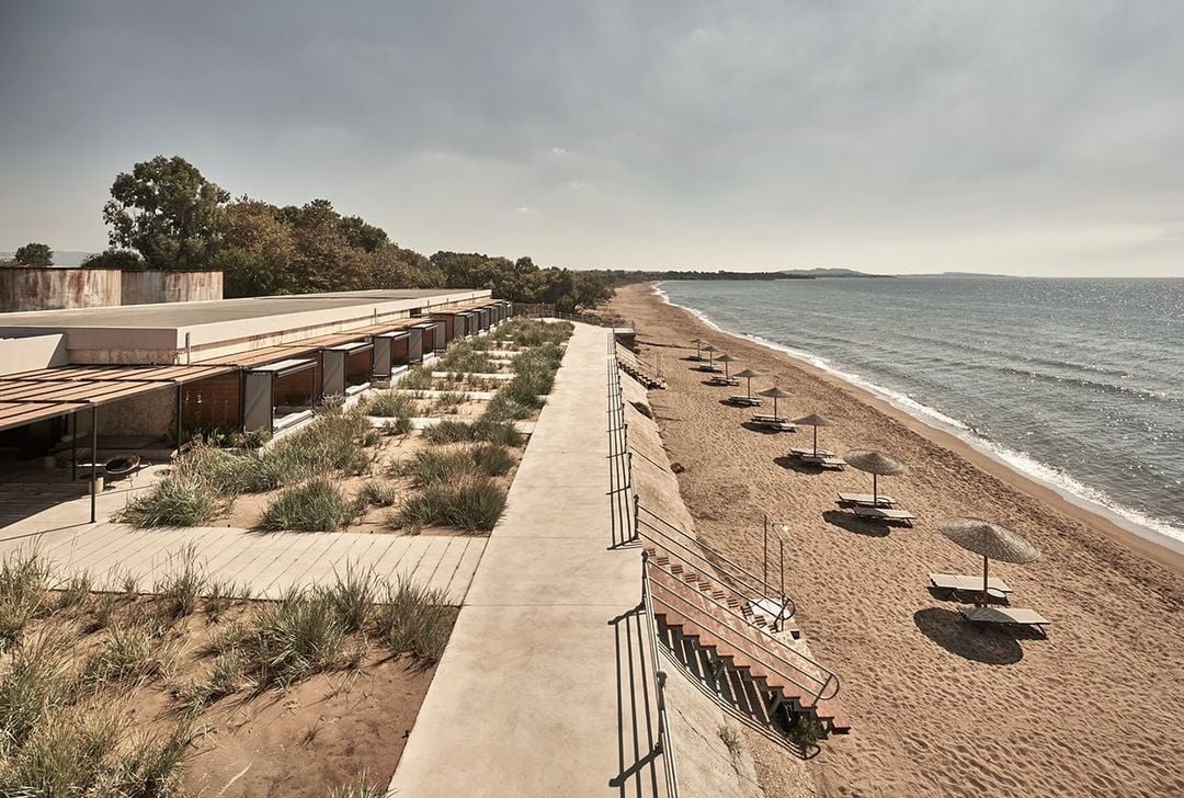 Dexamenes Seaside Hotel, Europe — Modern concrete pavilion with weathered timber screens overlooking sandy beach, native plantings, and calm Mediterranean waters under overcast sky.