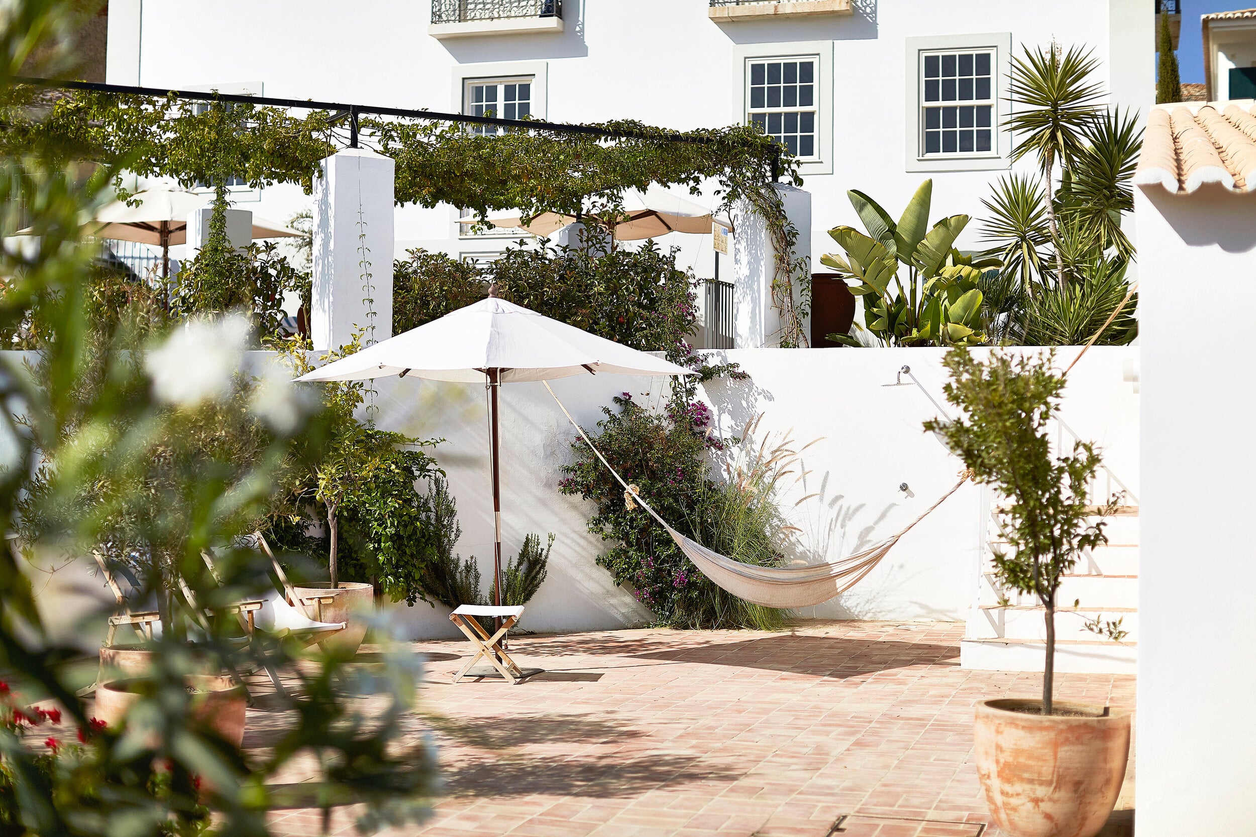 Casa Mãe, Europe — Courtyard with white umbrella, hammock, potted plants, and ivy-draped walls under Mediterranean sunlight.