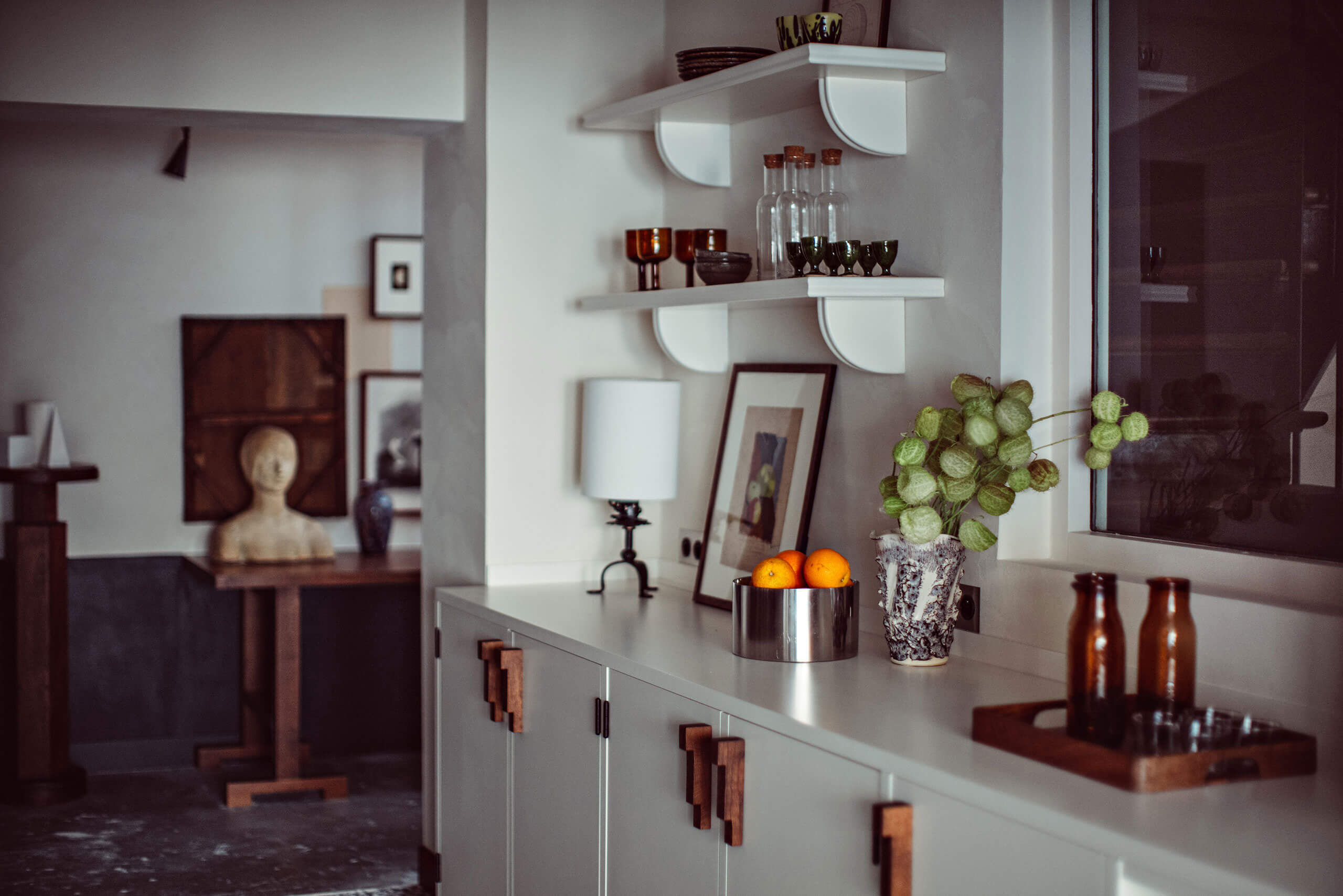 Académies et des Arts, Europe — Minimalist kitchen display with white cabinetry, wooden handles, floating shelves, potted plants, and decorative vessels in warm earth tones.