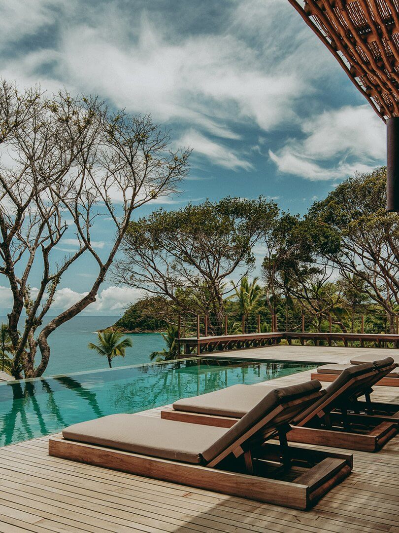 Barracuda Hotel & Villas, South America — Infinity pool with teak loungers overlooking turquoise ocean, sculptural trees, wooden deck and railing against clouded sky.