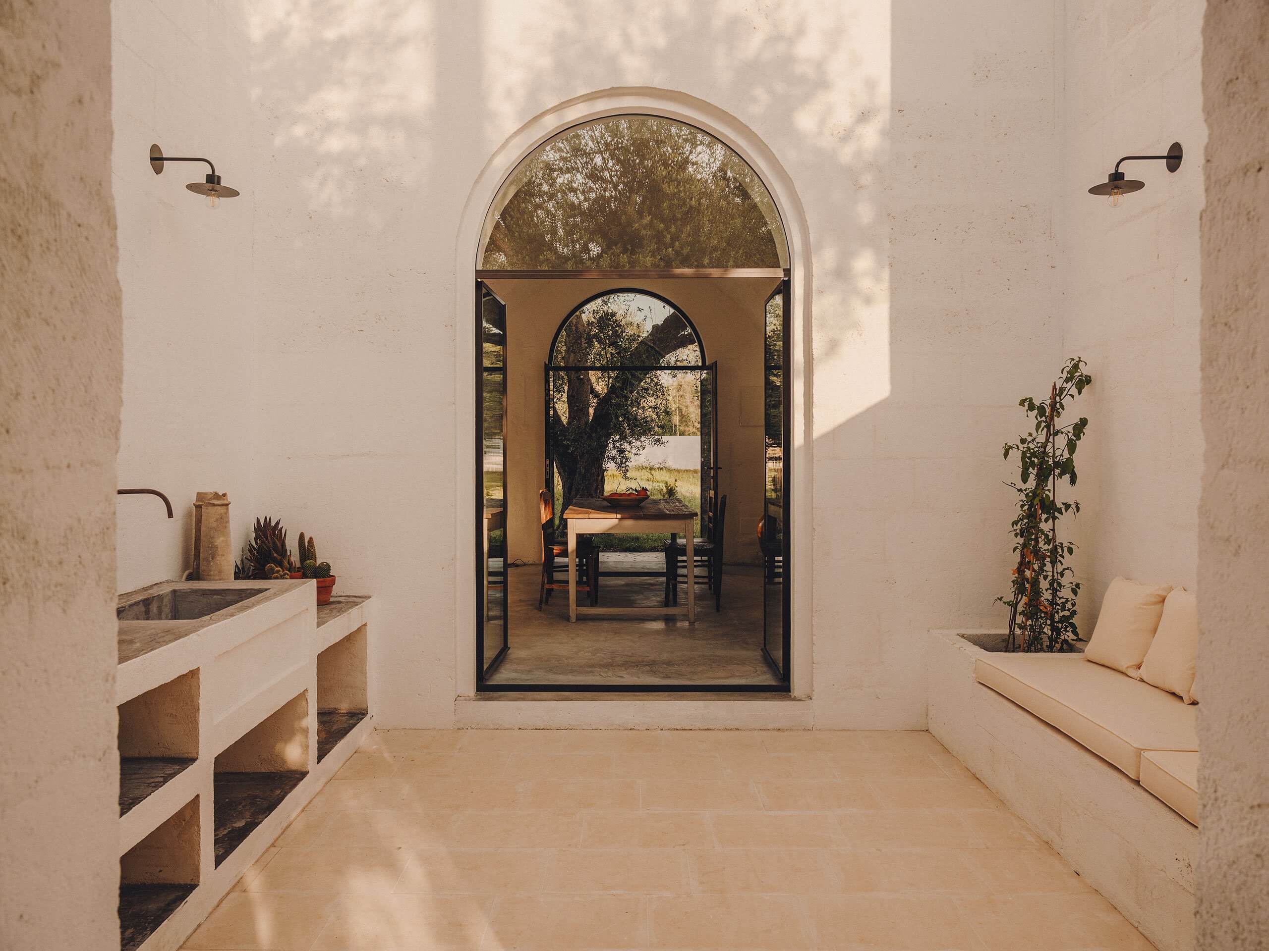 Borgo Gallana, Europe — Arched doorway with black steel frame opens to terracotta courtyard, cream stone walls, built-in bench seating, potted succulents, morning light.
