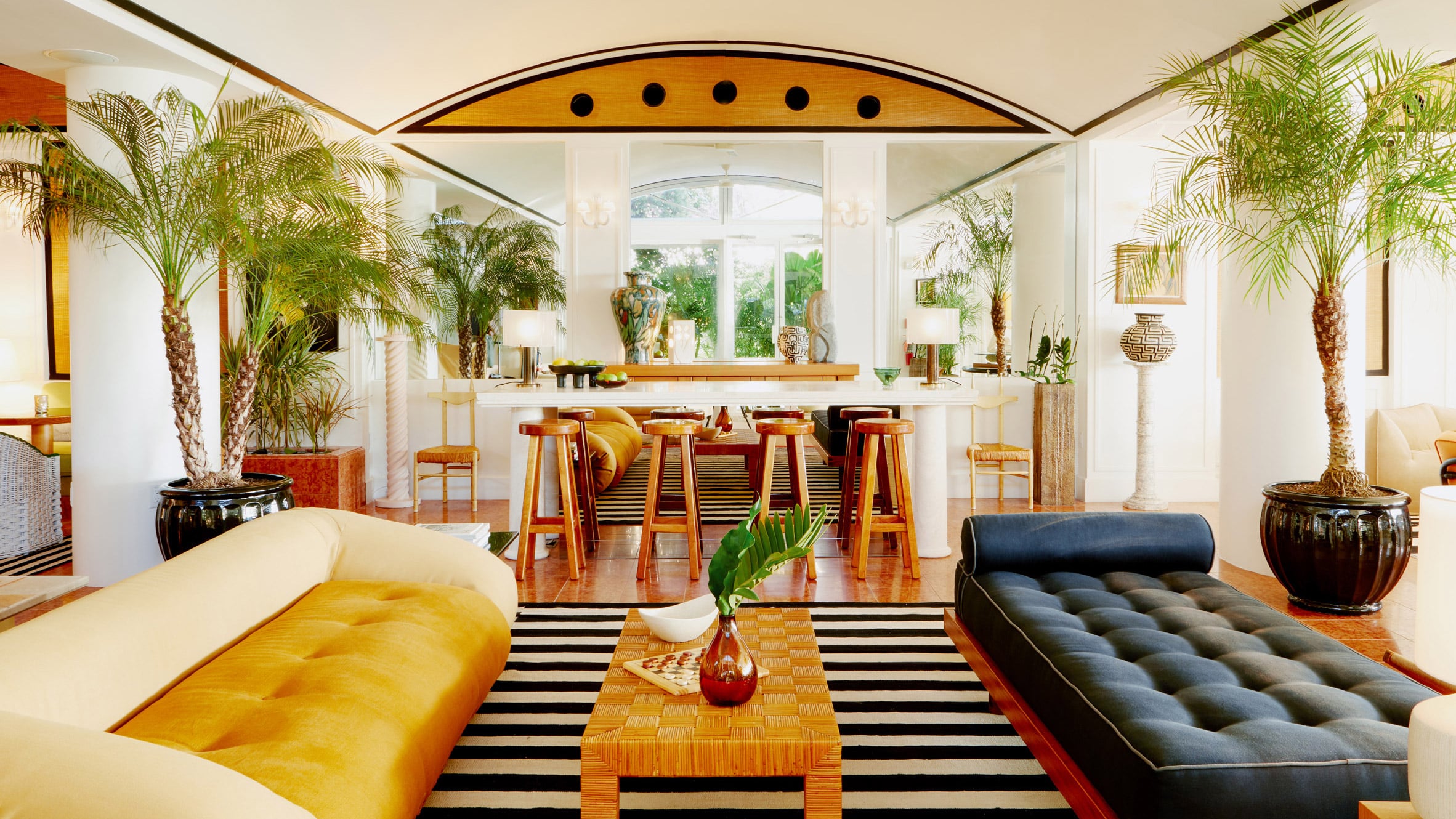 Palm Heights, North America — Mid-century modern lounge with golden velvet seating, black-and-white striped rug, potted palms, wooden bar stools, and geometric ceiling detail.