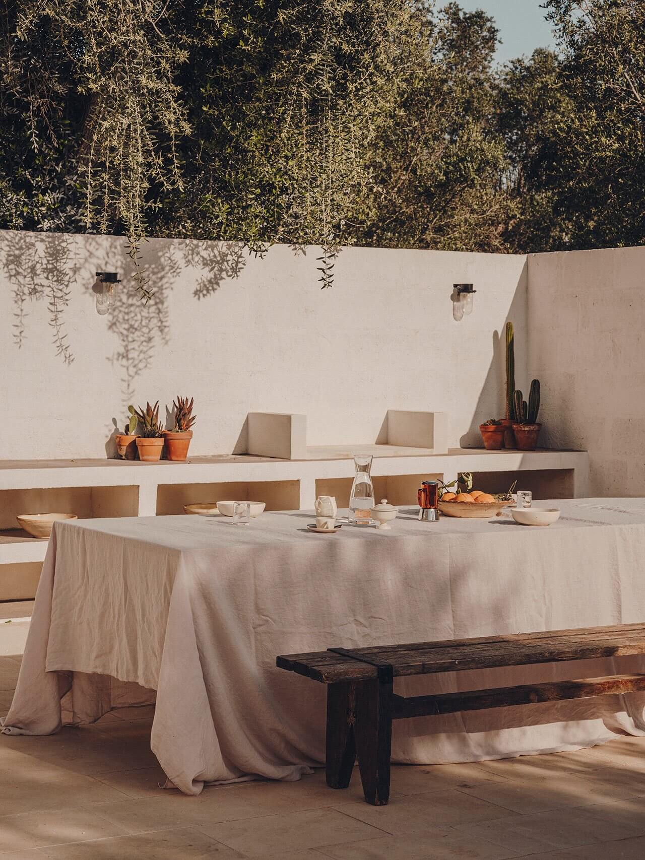 Borgo Gallana, Europe — Minimalist outdoor dining terrace with cream linen tablecloth, dark wood bench, potted succulents, and cascading ivy against whitewashed concrete walls.