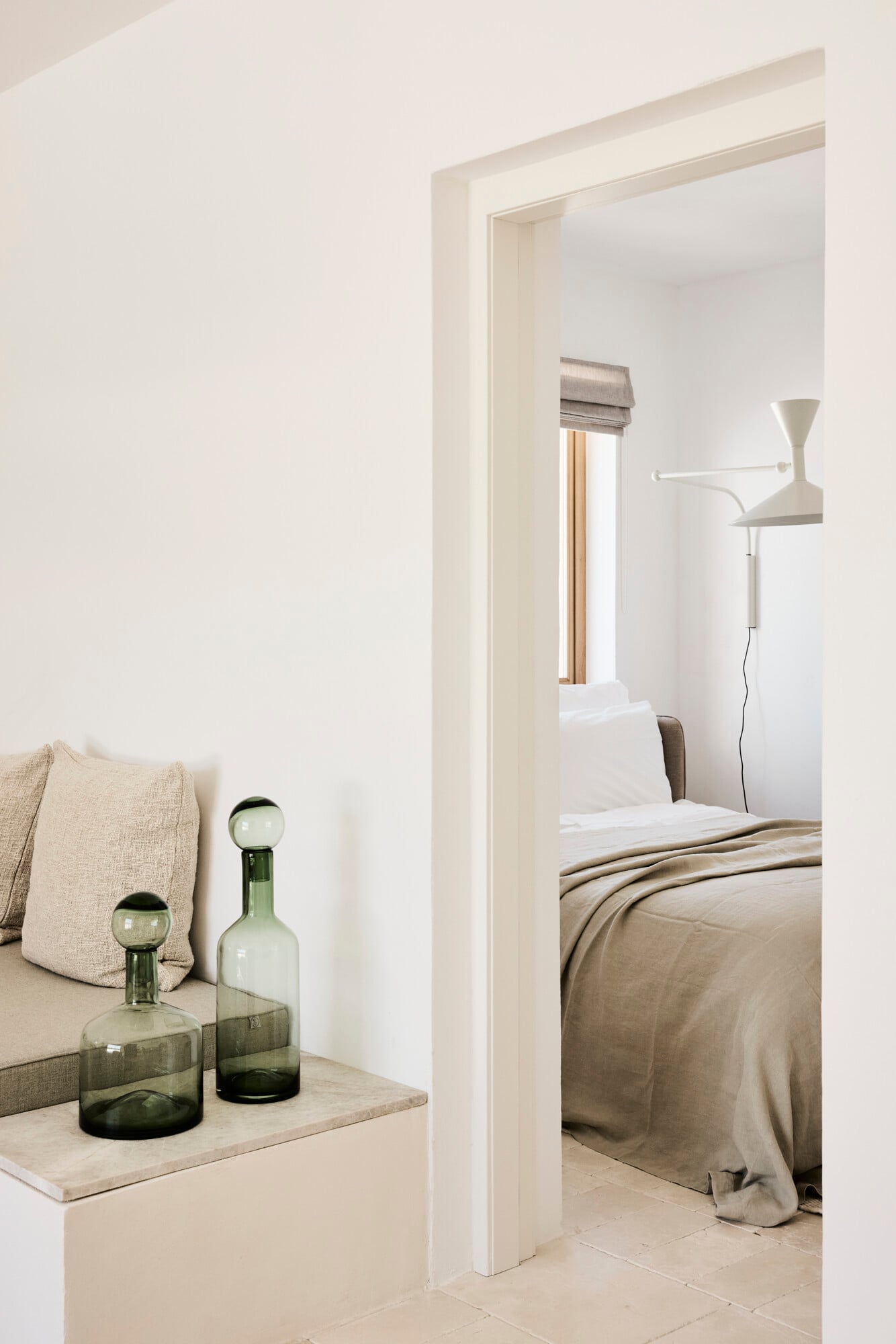 Villa Sessana, Italy — Minimalist bedroom suite with neutral linen furnishings, white stone archway, glass vessels, and natural light from window with roman shade.