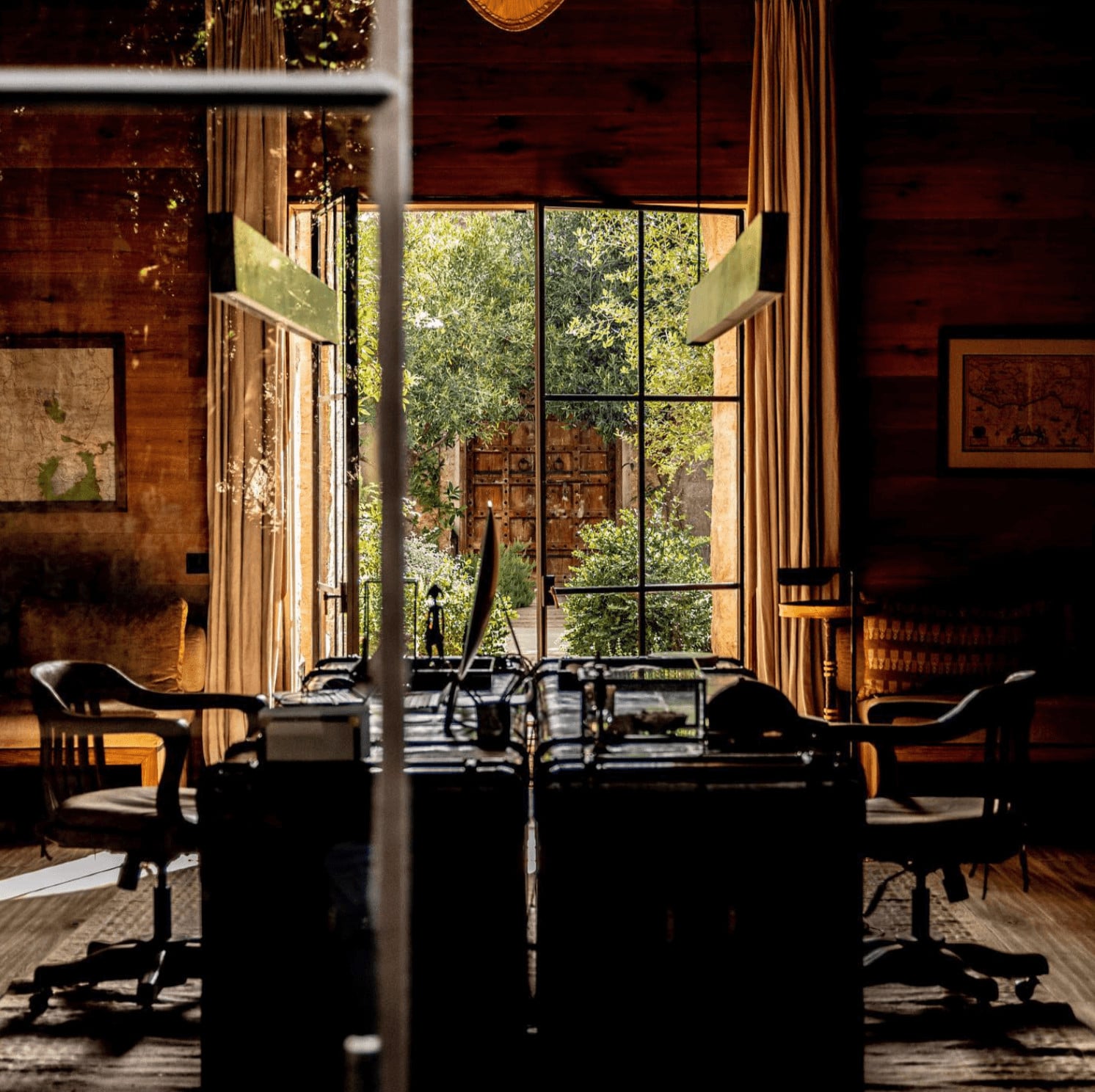 Arijiju Retreat, Africa — Sunlit interior with floor-to-ceiling windows framing lush vegetation, wooden furnishings, golden curtains, and ivy-covered courtyard beyond.
