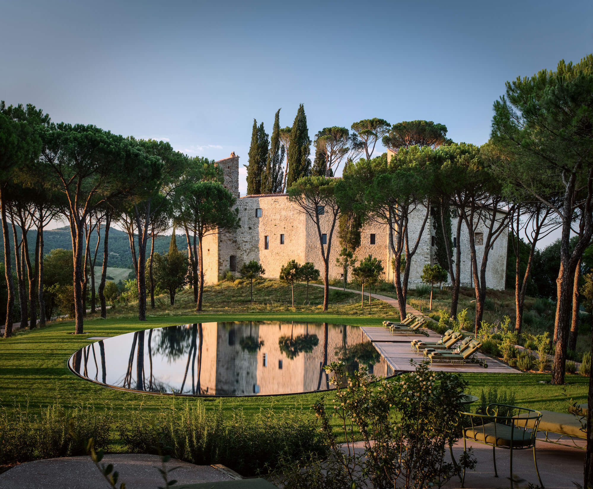 Castello di Reschio, Umbria, Italy — Medieval stone tower reflected in modernist pool surrounded by Italian cypress trees and manicured grounds at golden hour.