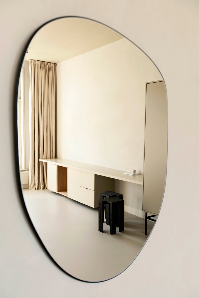 Hotel âme, Europe — Oval mirror reflecting minimalist bedroom with cream desk, black nesting tables, floor-to-ceiling curtains, and soft natural light.