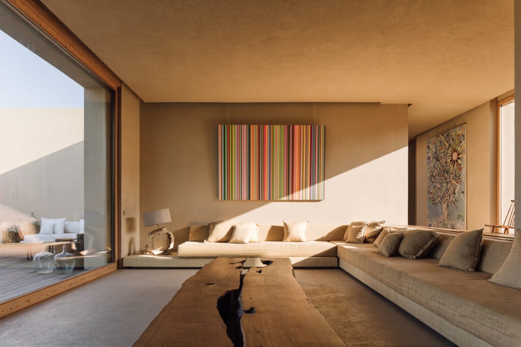 Melides Art, Portugal — Minimalist living room with sunlit concrete floors, built-in seating, natural wood table, and colorful striped artwork on warm ochre walls.