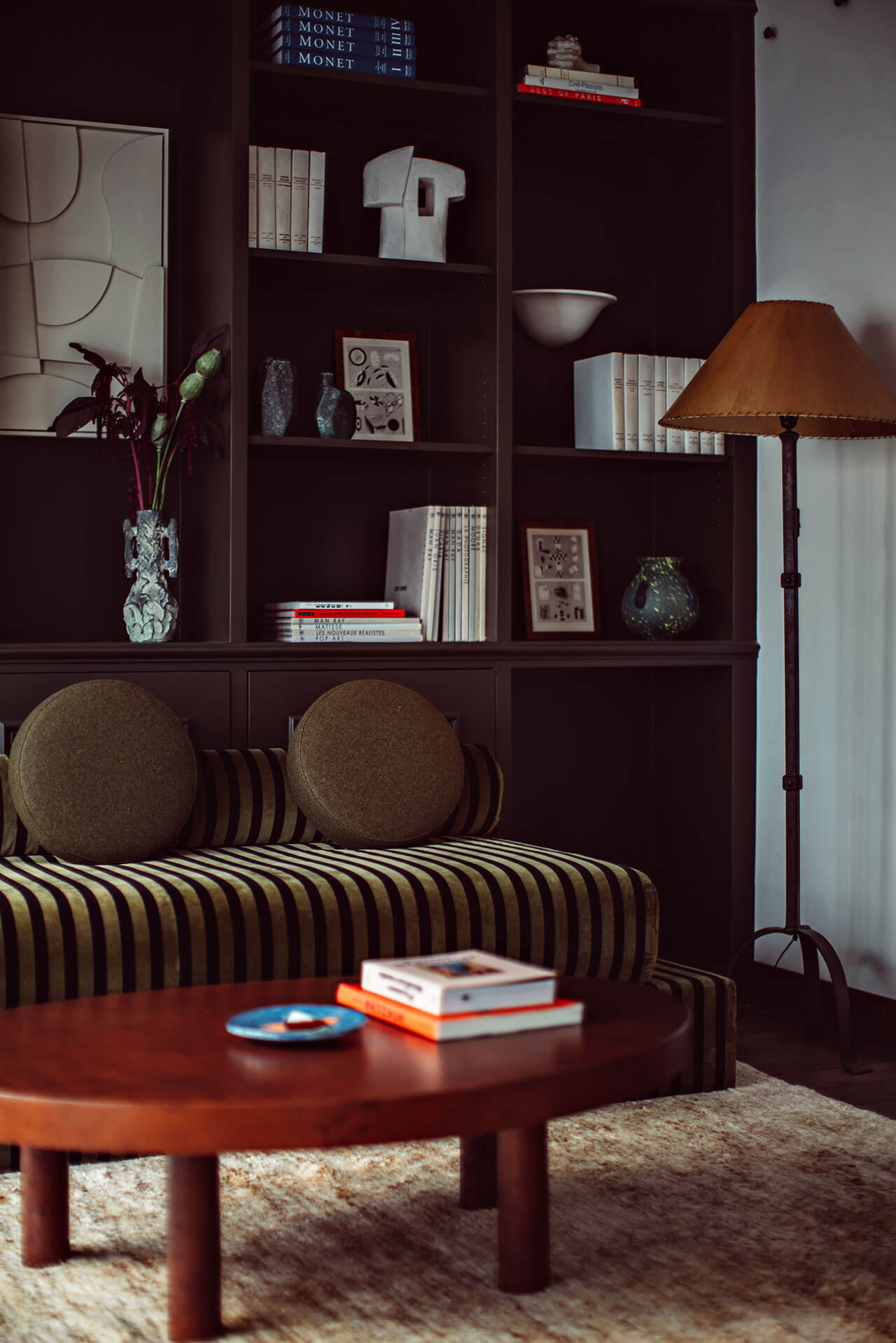 Académies et des Arts, Europe — Dark wood shelving with curated books and ceramics, striped sofa with round pillows, warm leather floor lamp, and round wood table with magazines on neutral rug.