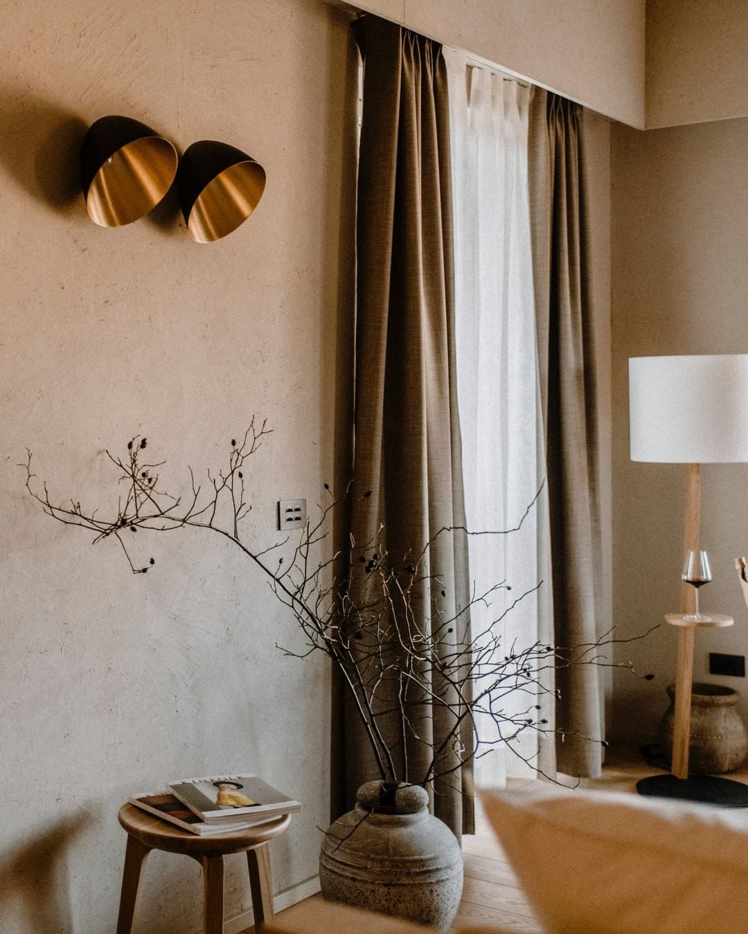Casa di Langa, Europe — Minimalist bedroom alcove with layered linen curtains, dried botanicals in terracotta vessel, brass wall sconces, and wooden side table with magazines.