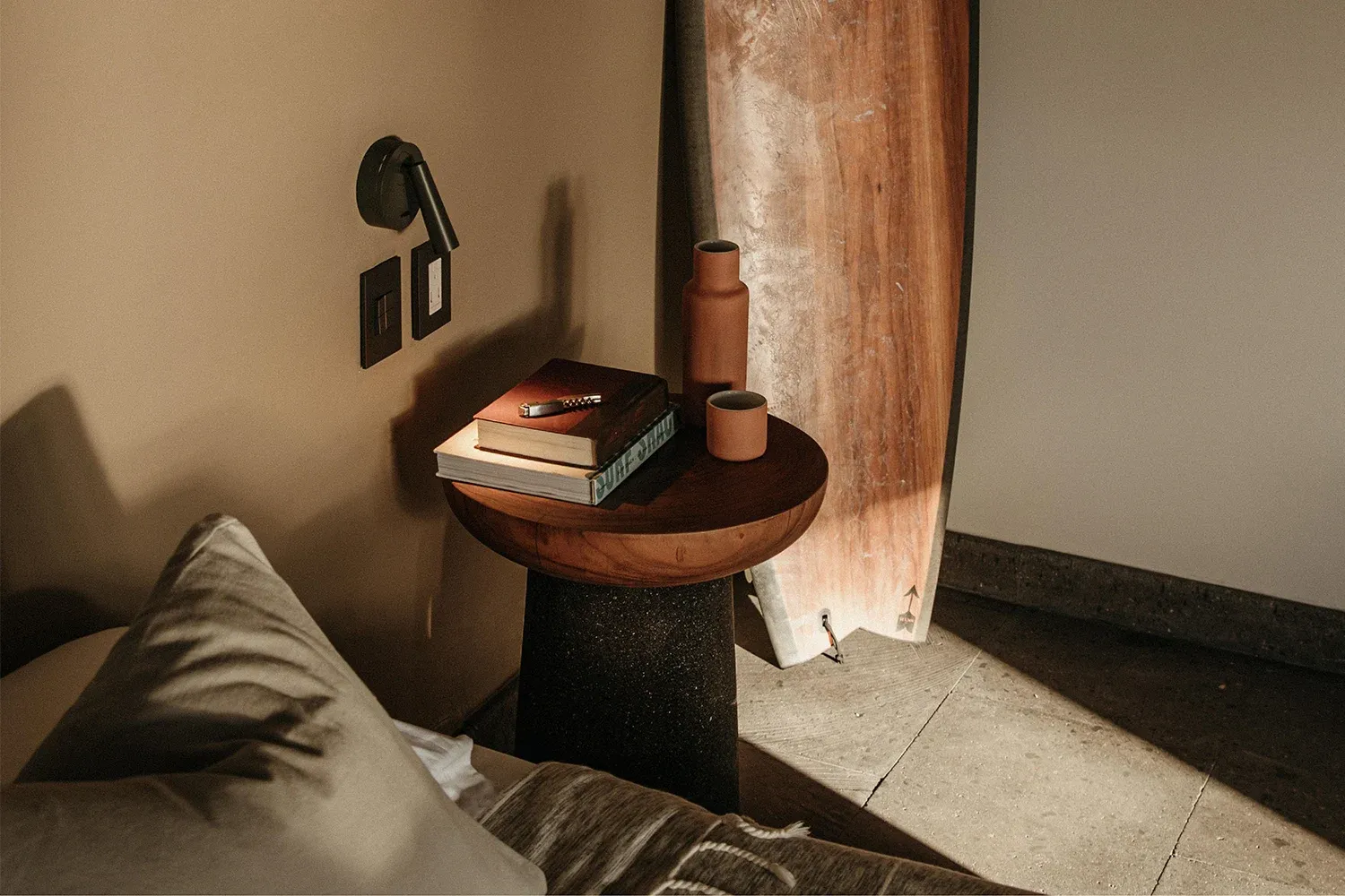 Hotelito MUSA, Mexico — Minimalist bedroom corner with wooden side table, stacked books, terracotta ceramics, and natural light casting shadows on neutral walls.