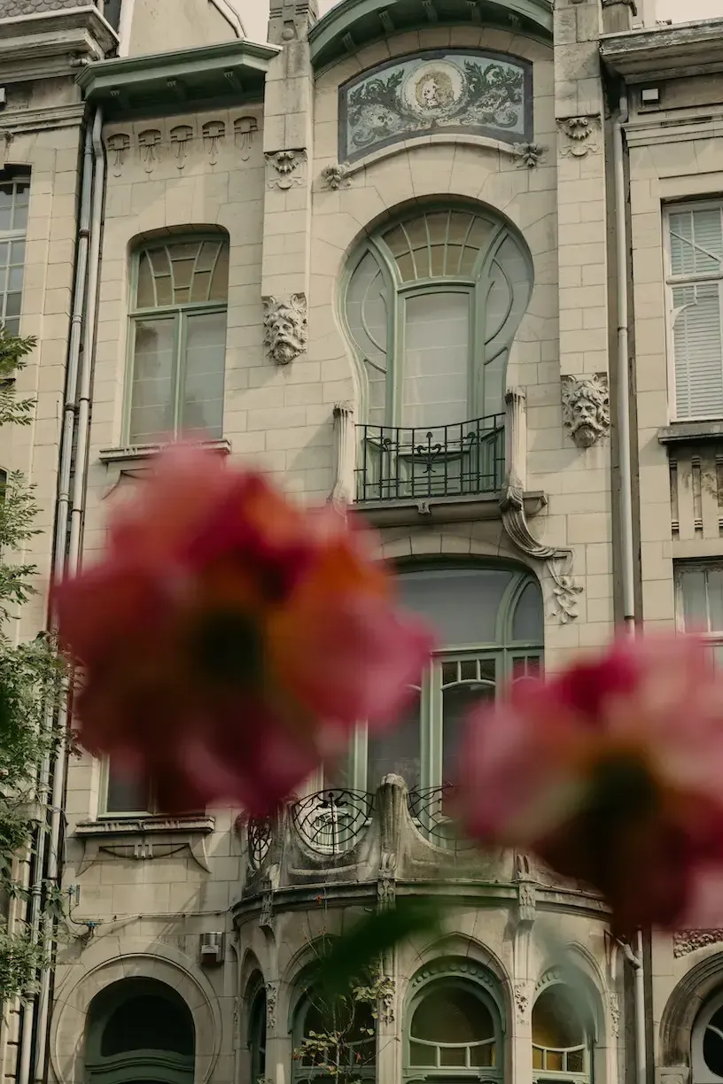August, Europe — Pale stone facade with arched windows, decorative lion masks, and wrought-iron balconies framed by blurred crimson roses in foreground.