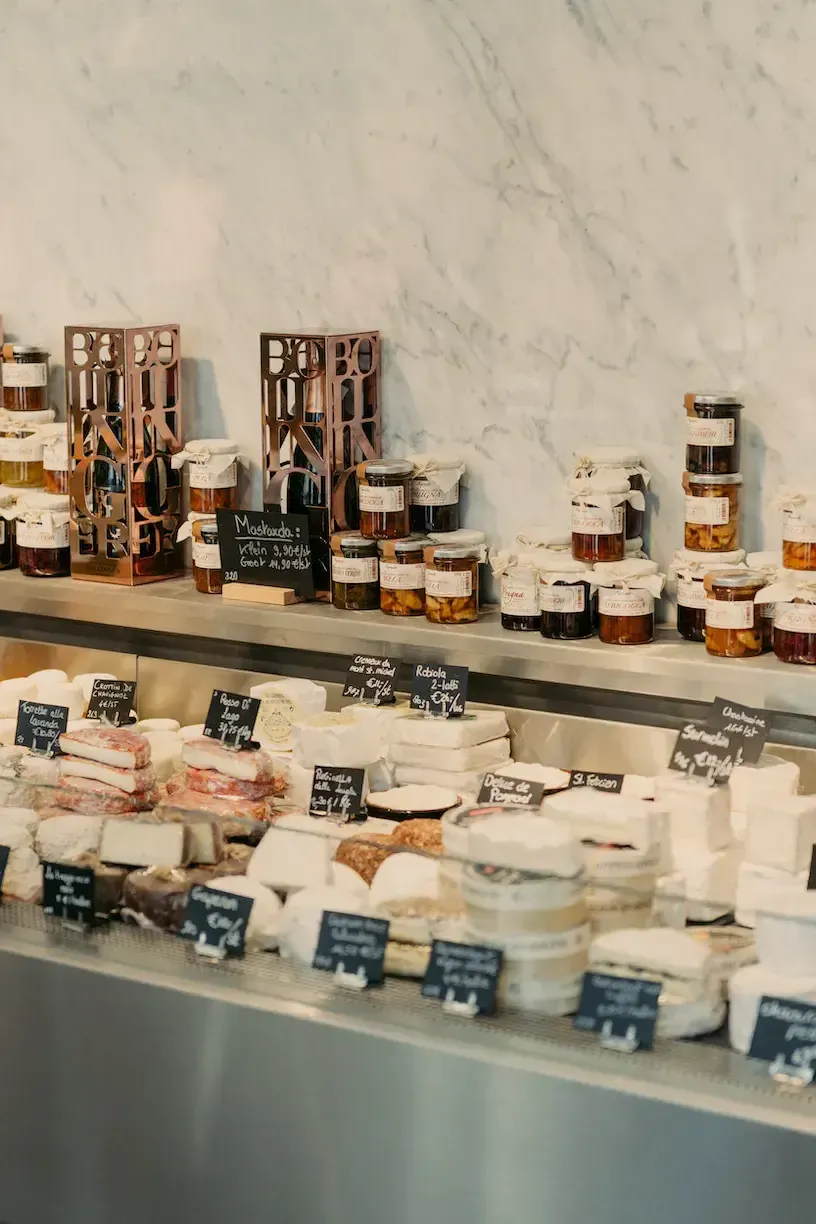 Only Cheese, Europe — Marble-backed delicatessen counter displaying artisanal cheeses, cured meats, and preserves in glass jars with handwritten labels, warm natural light.
