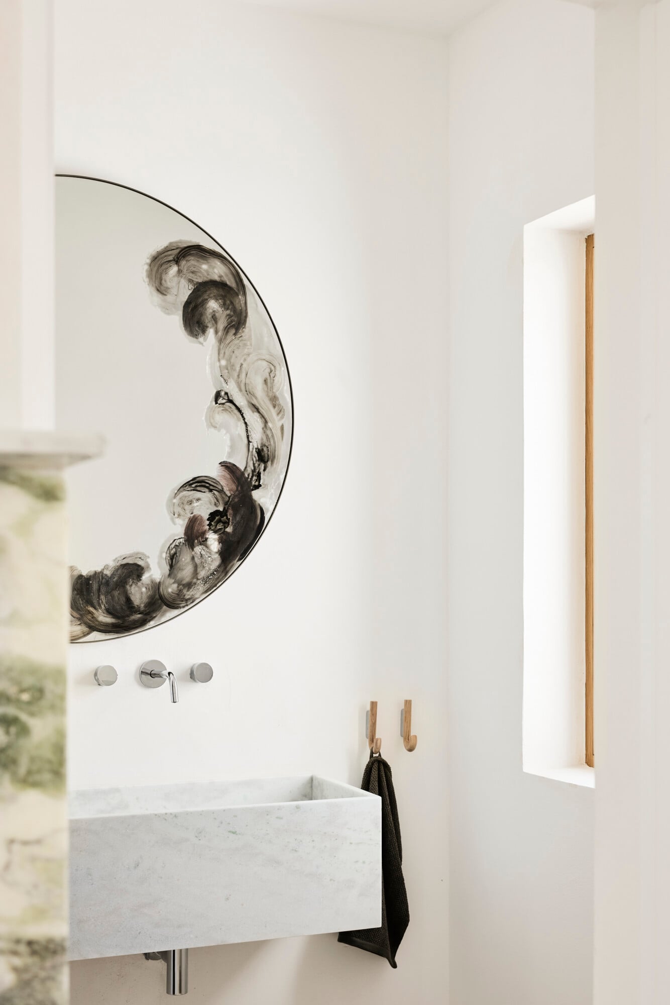 Villa Sessana, Italy — Minimalist bathroom with curved marble sink, sculptural mirror with organic ink-wash artistry, wooden fixtures, and natural light from recessed window.