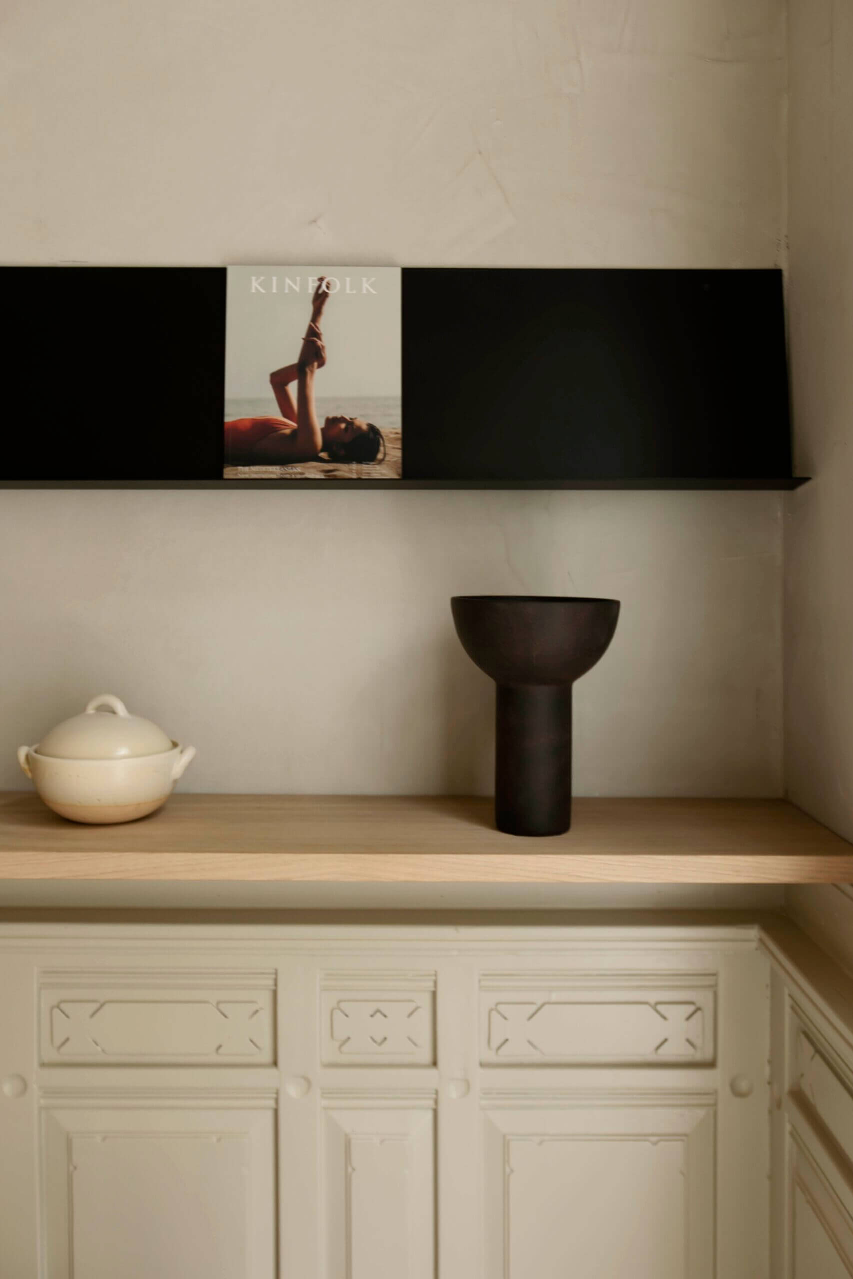Hotel âme, Europe — Minimalist shelf styling with cream ceramic vessel, black sculptural vase, and framed portrait above natural wood surface against cream wall.