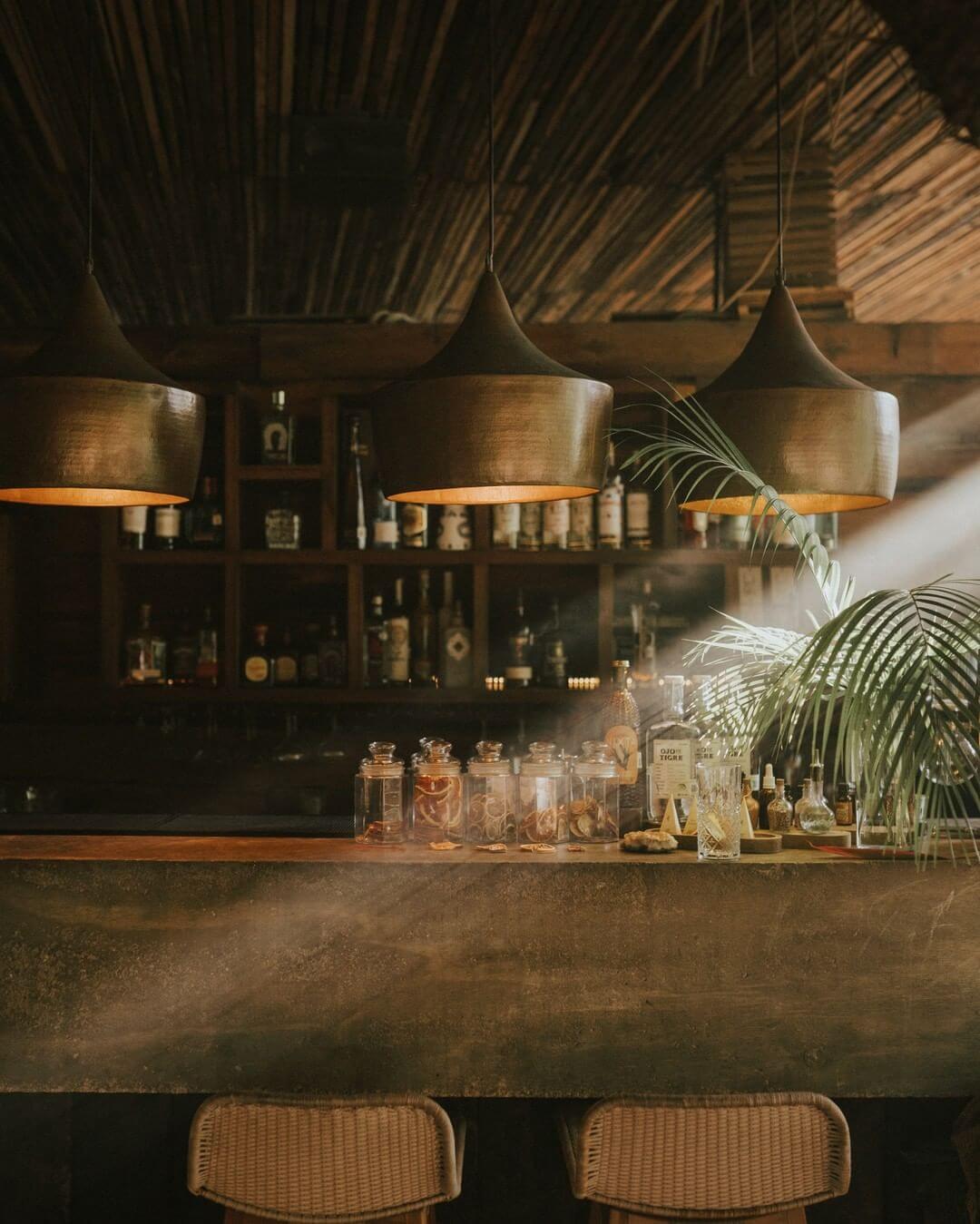 Be Tulum, Mexico — Sunlit bar counter with metallic pendant lights, woven ceiling, concrete surface, glass bottles, and potted palm plant.