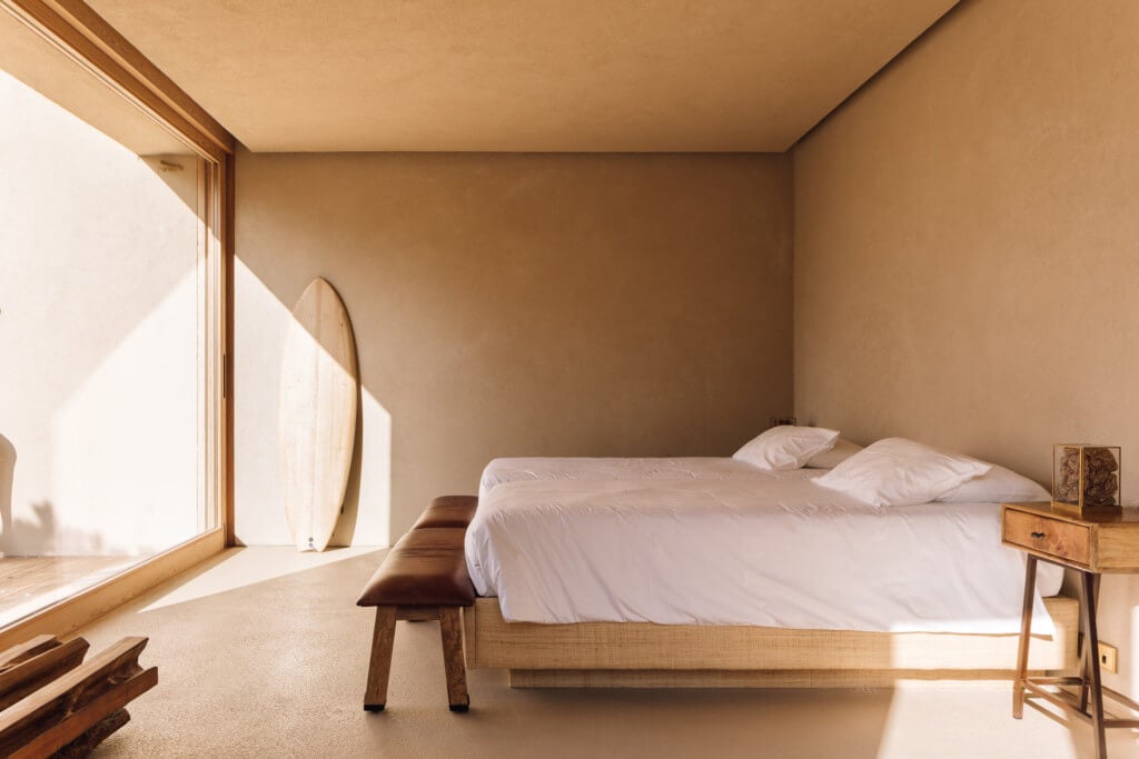 Melides Art, Portugal — Minimalist bedroom with natural light, wooden bed frame, cream bedding, terracotta walls, and modernist furnishings.