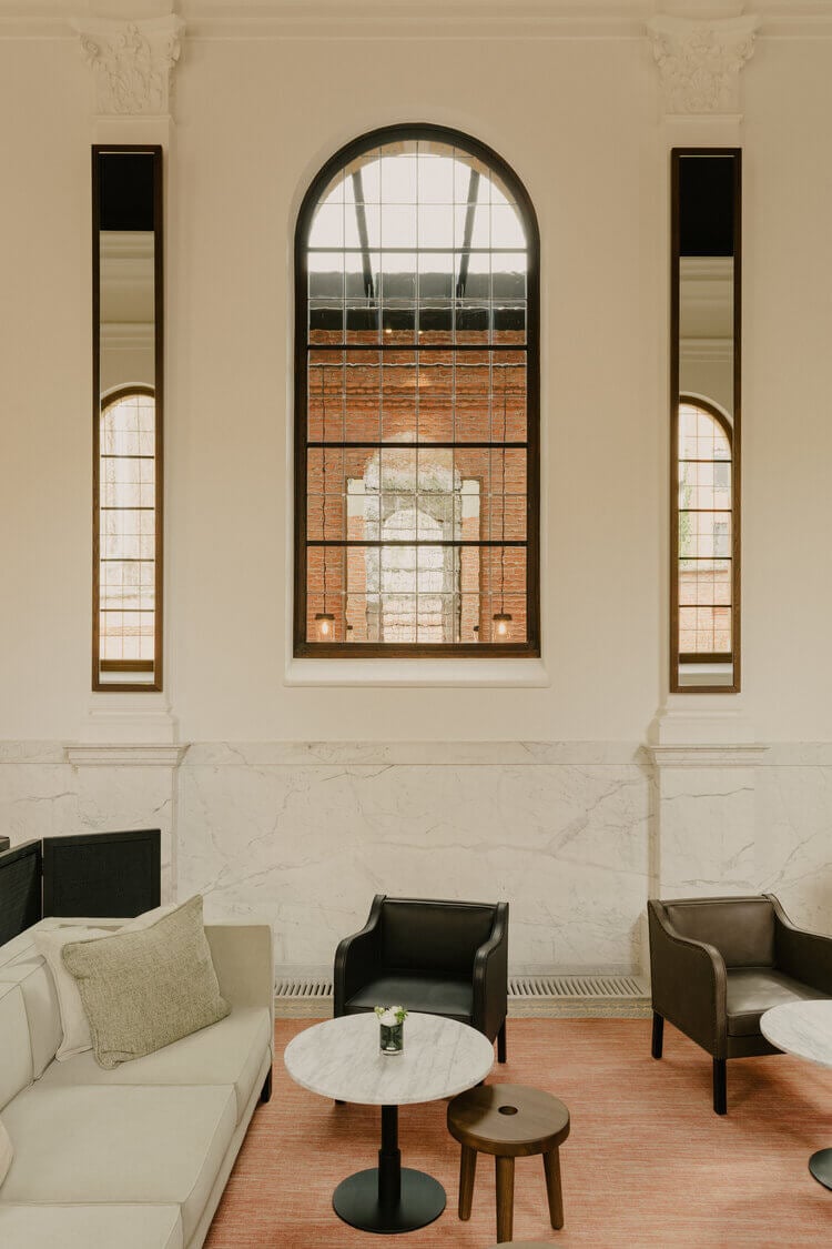 August, Europe — Minimalist lounge with cream sofa, black leather chairs, arched windows overlooking brick building, marble walls, wooden floors, decorative corbels.