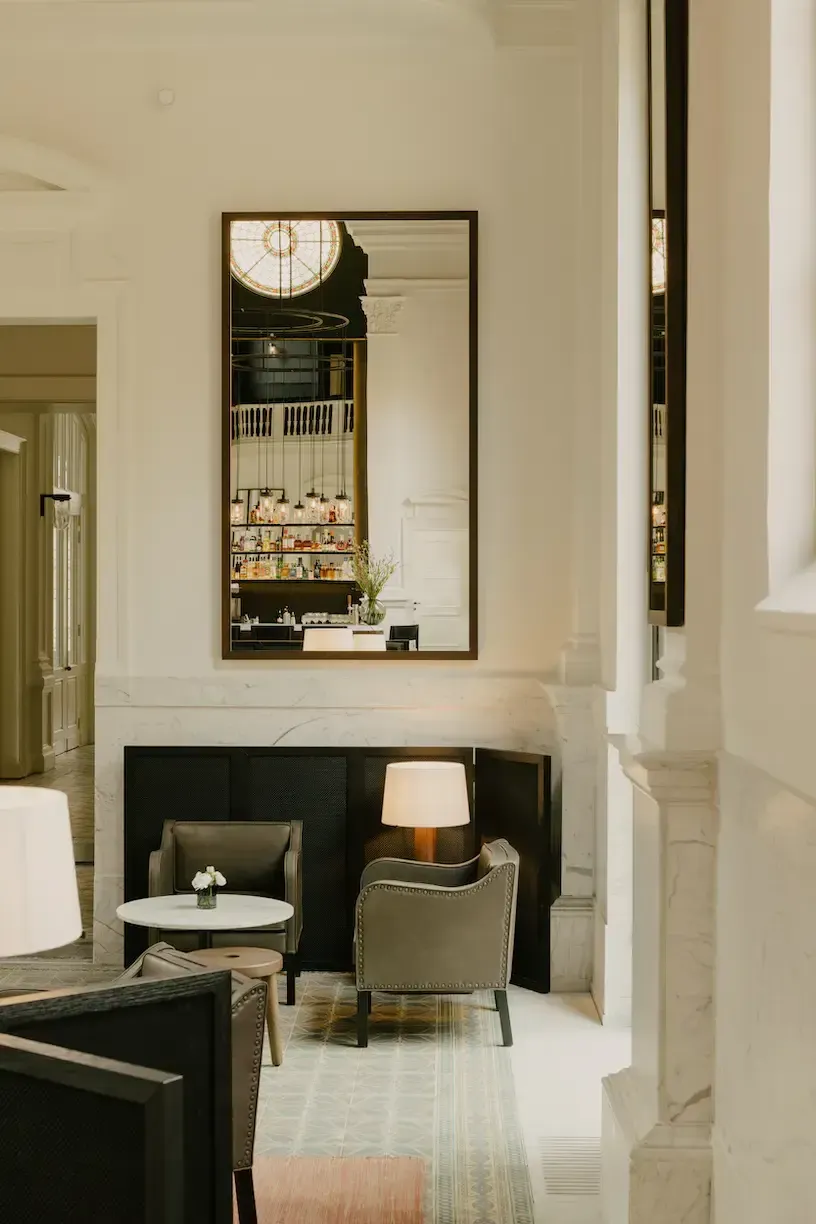 August, Europe — Modern hotel corridor with marble flooring, mirrored wall panel, tailored seating nook with upholstered chairs, brass lamp, and minimalist neutral palette.