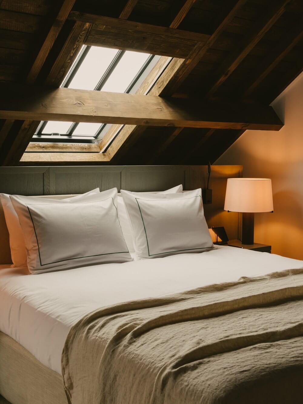 August, Europe — Attic bedroom with skylights, wooden beams, white linens, olive throw, brass lamp, warm ochre walls, and natural daylight.