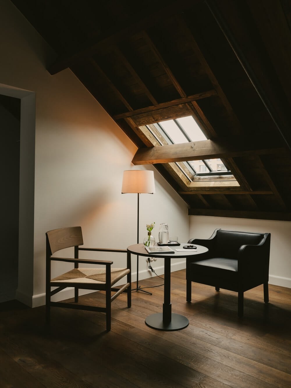 August, Europe — Attic room with exposed wooden beams, skylights, minimalist seating, warm natural light on white walls, blonde wood flooring, contemporary hospitality design.