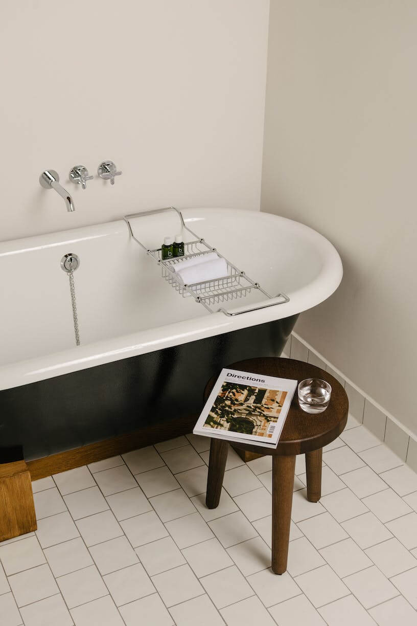 August, Europe — Minimalist bathroom with freestanding black-and-white soiled tub, chrome fixtures, wooden side table, and white tile flooring.