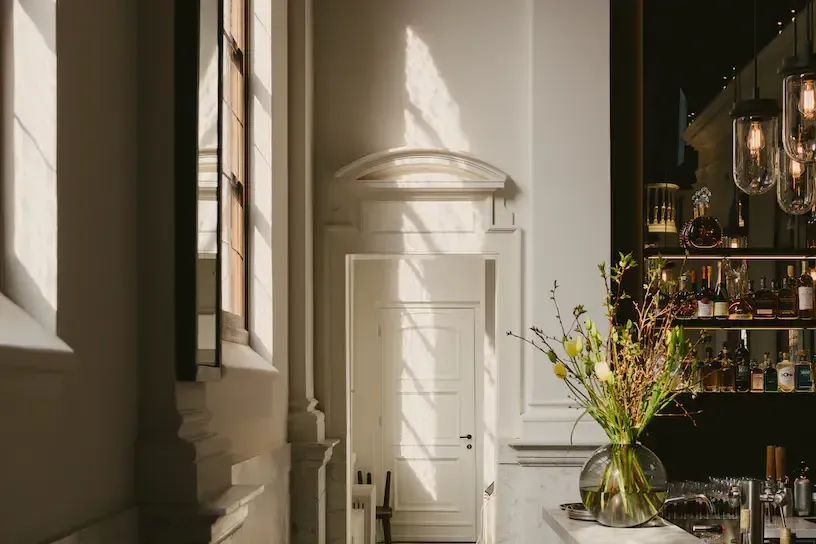 August, Europe — Sunlit corridor with cream paneled door, arched transom window, fresh flowers in glass vase, luxury hospitality design interior.