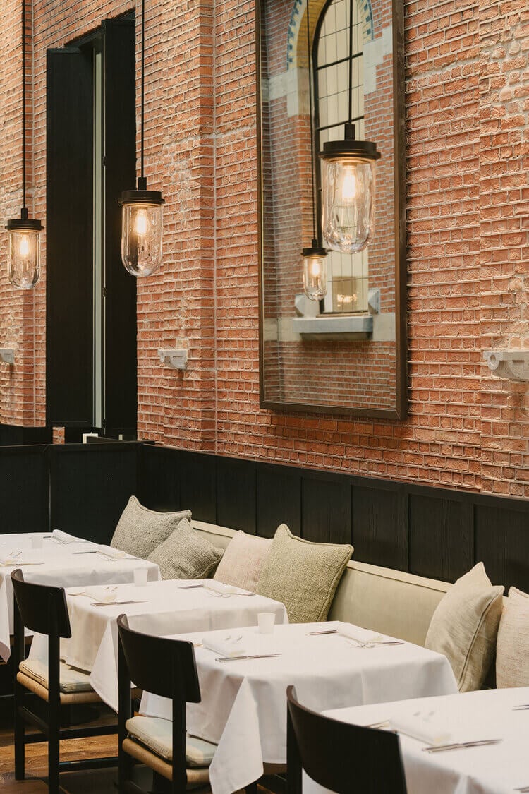 August Restaurant, Europe — Exposed brick dining room with black banquette seating, pendant lights, mirror, and white linens creating intimate industrial-modern ambiance.