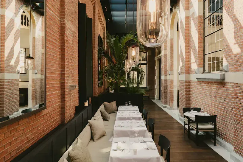 August, Europe — Narrow brick dining corridor with long banquette seating, skylight, potted palm, and warm evening ambient lighting.
