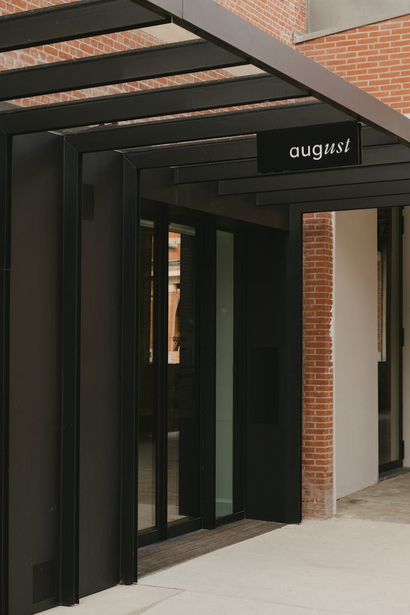 August, Europe — Minimalist black storefront with glass doors and steel pergola canopy against exposed brick, modern hospitality design detail.