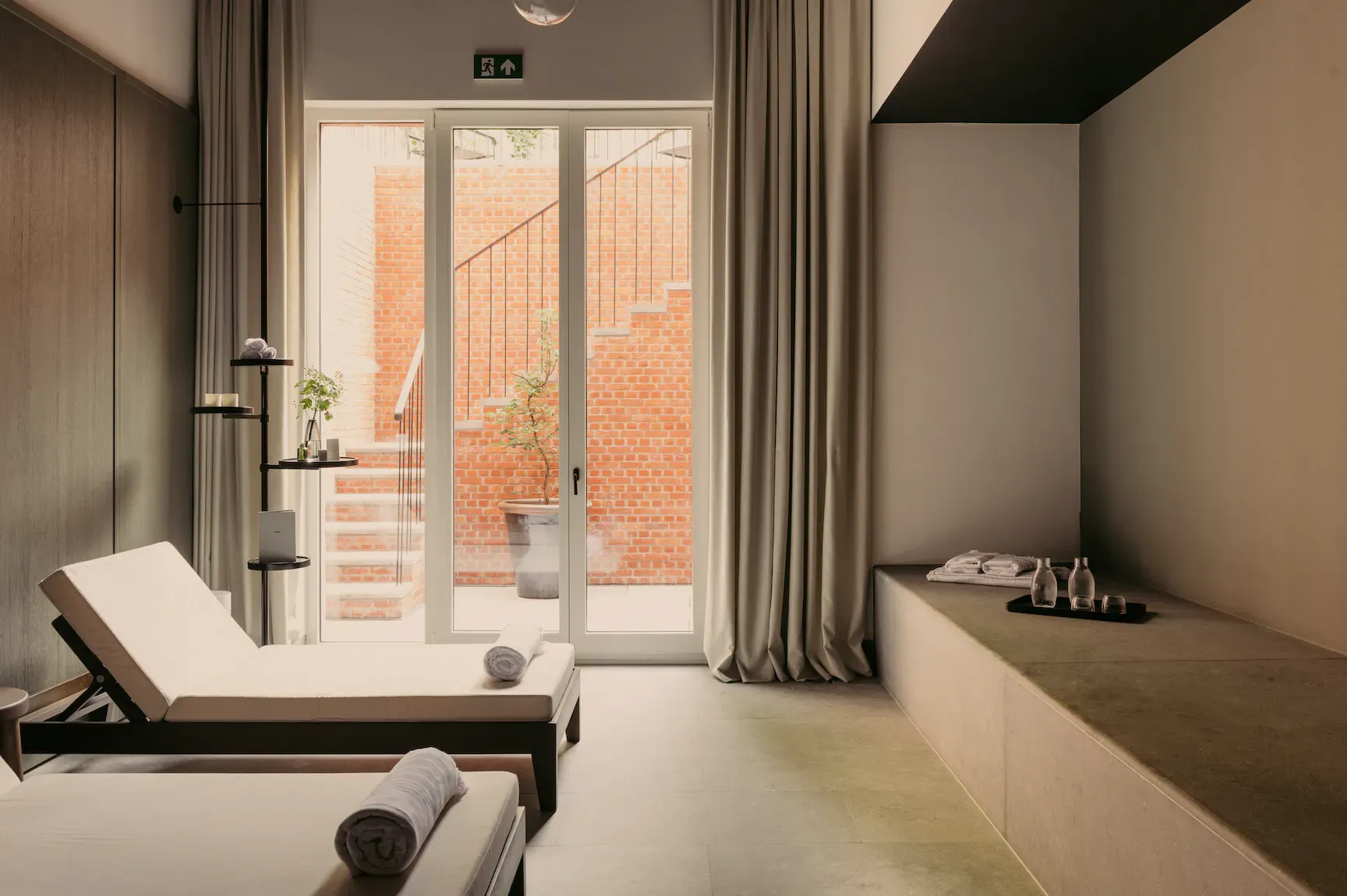 August Spa, Europe — Minimalist spa relaxation room with cream loungers, brick courtyard view, natural light, and neutral palette.