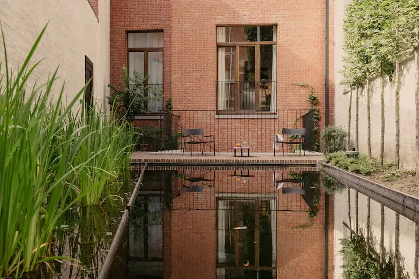 August, Europe — Brick courtyard with reflecting pool, minimalist seating, ivy-covered walls, and large windows overlooking serene architectural space.