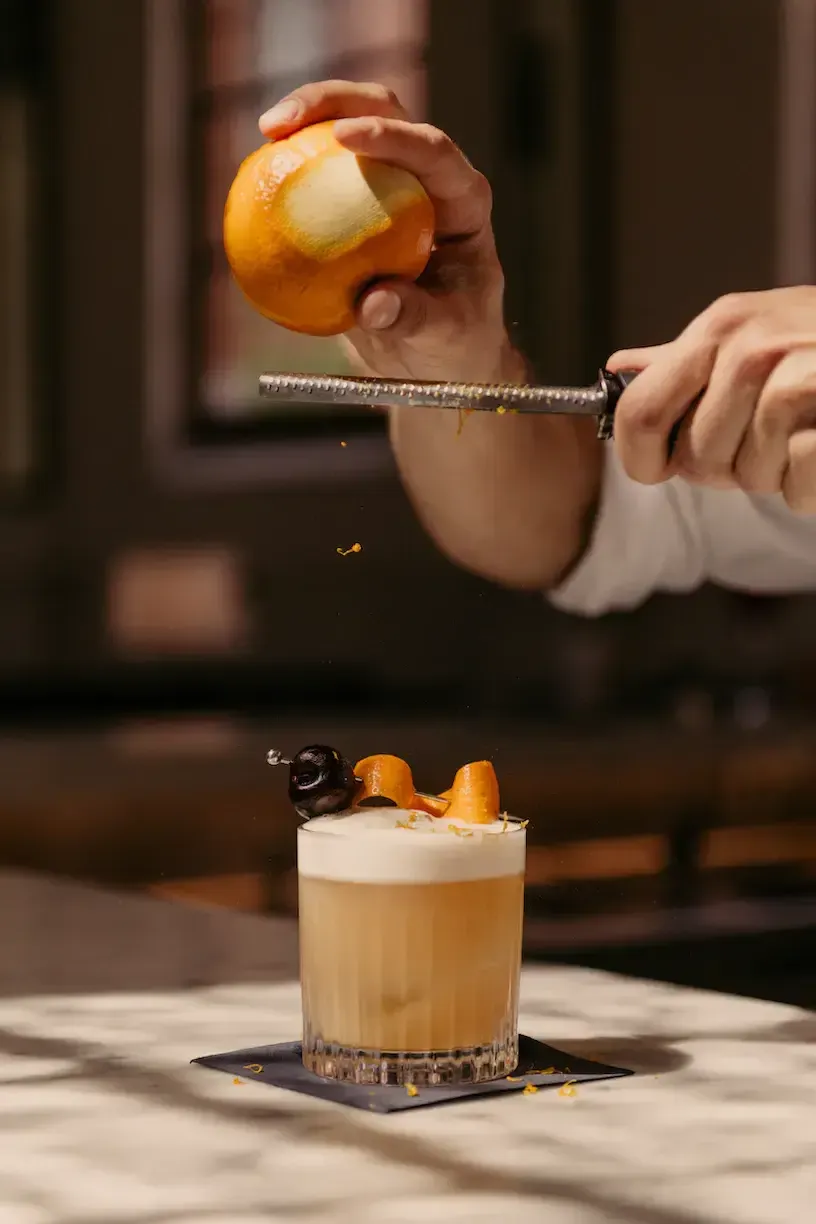 August — Bartender zesting orange peel over craft cocktail with microplane, amber liquid and foam, luxury hotel bar ambiance.