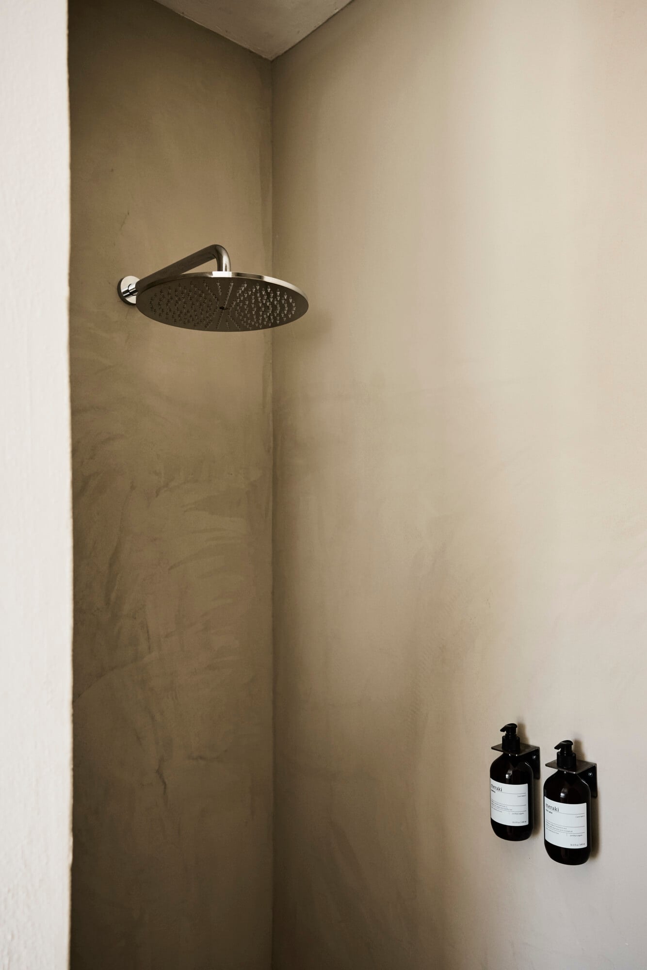 Villa Sessana, Italy — Minimalist shower niche with black rainfall showerhead, concrete walls, and dark glass bottles on shelf against neutral plaster.