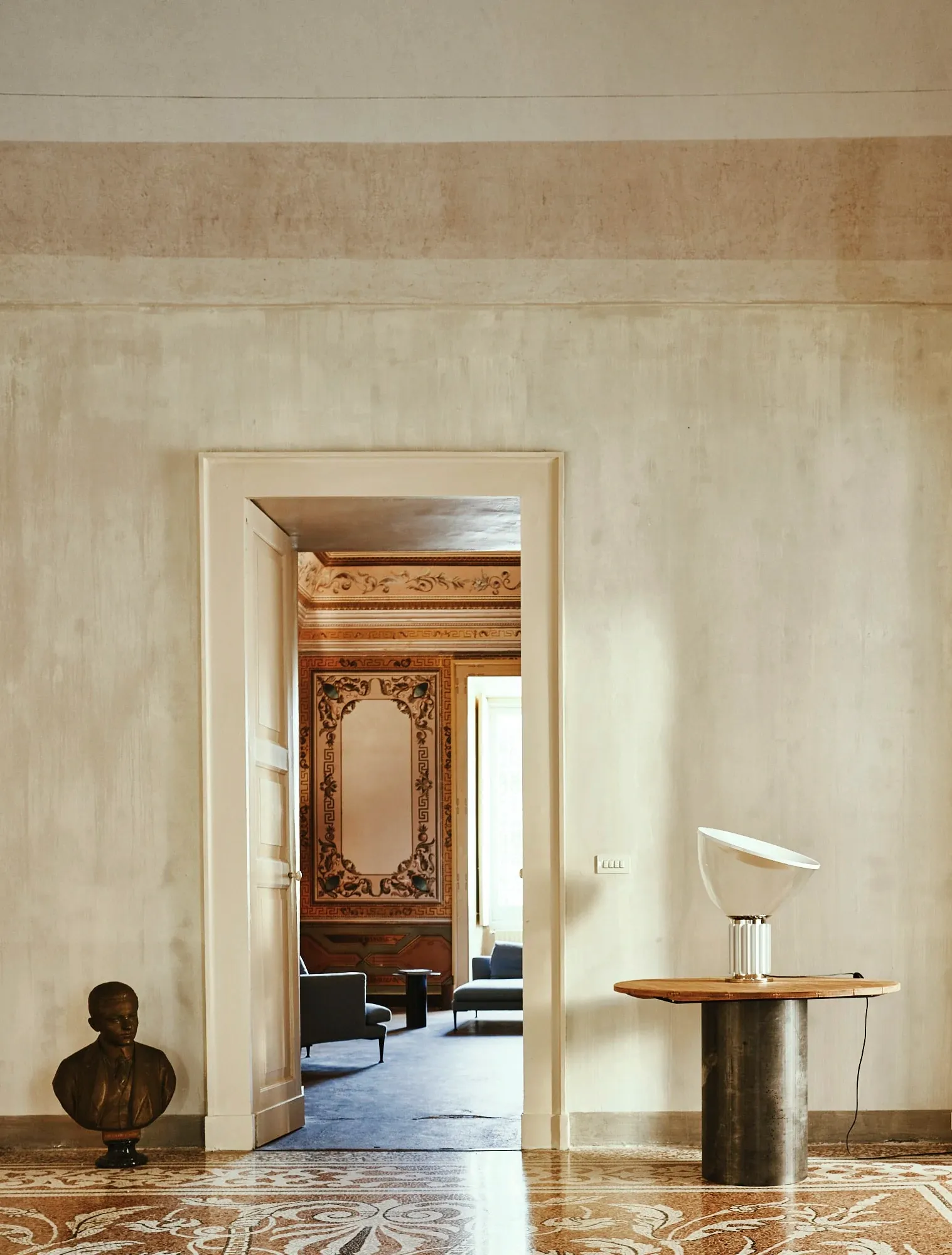 Palazzo Daniele, Europe — Neoclassical gallery with inlaid marble floors, ornate wooden door frame, bronze bust, and modernist table lamp beneath cream walls.