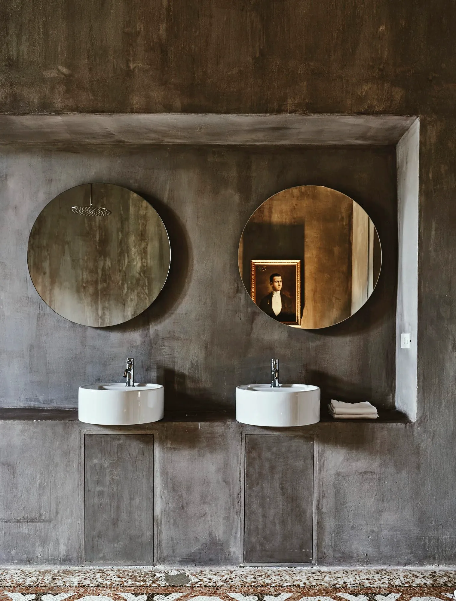 Palazzo Daniele, Europe — Dual circular mirrors above white vessel sinks set in weathered concrete vanity within minimalist brutalist bathroom.