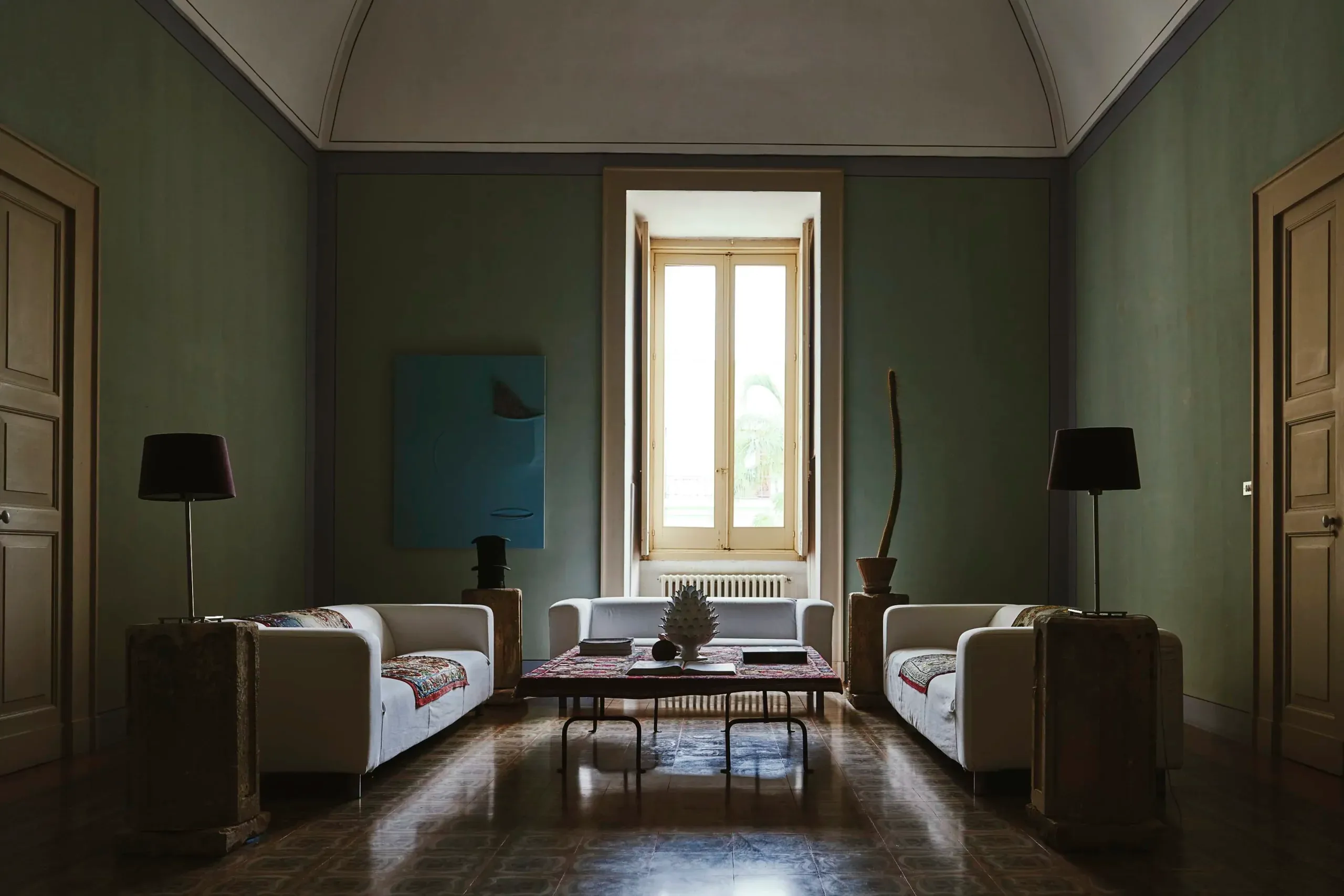 Palazzo Daniele, Europe — Symmetrical living room with sage green walls, cream sofas, dark floor lamps, blue artwork, and tall French doors opening to landscape light.