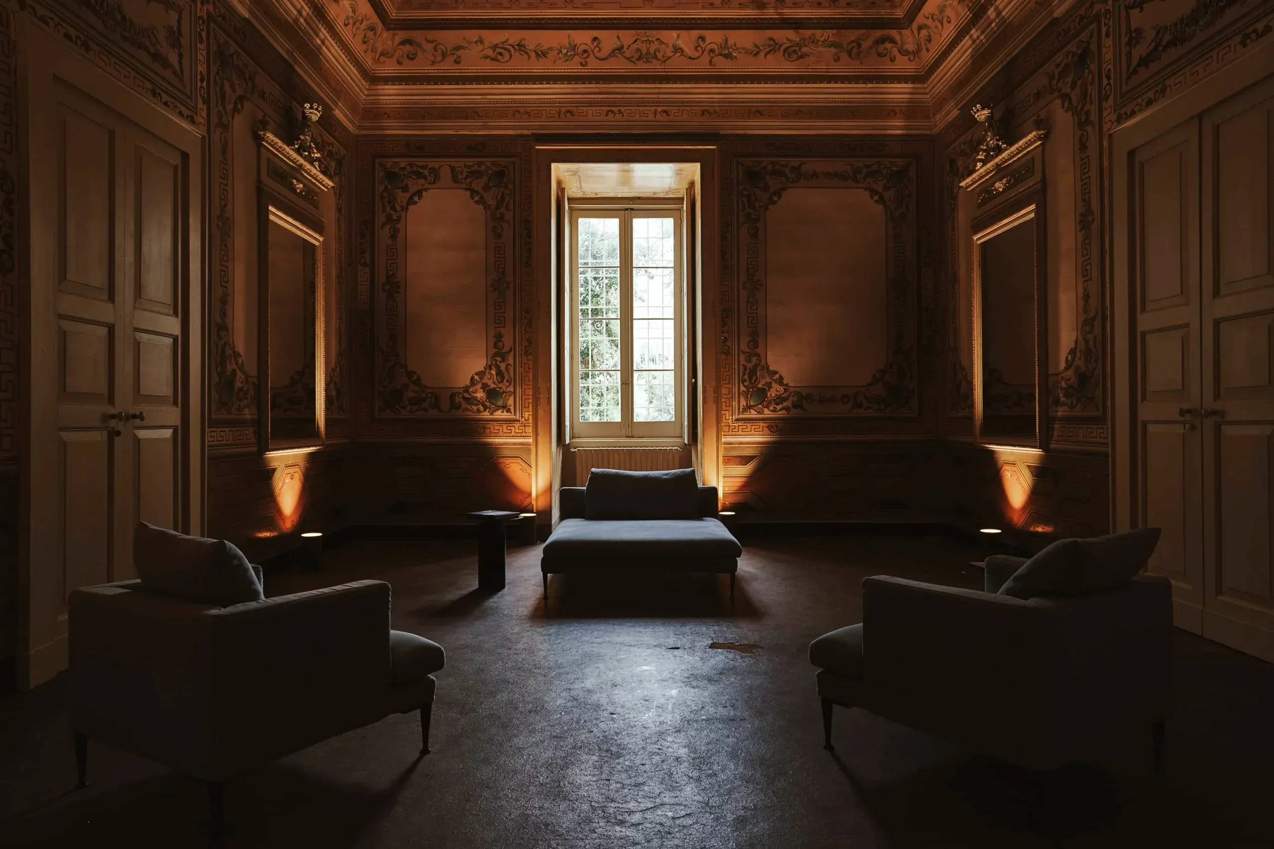 Palazzo Daniele, Europe — Neoclassical salon with gilded wall panels, ornate coffered ceiling, symmetrical seating, central bed, tall windows, warm ambient lighting.