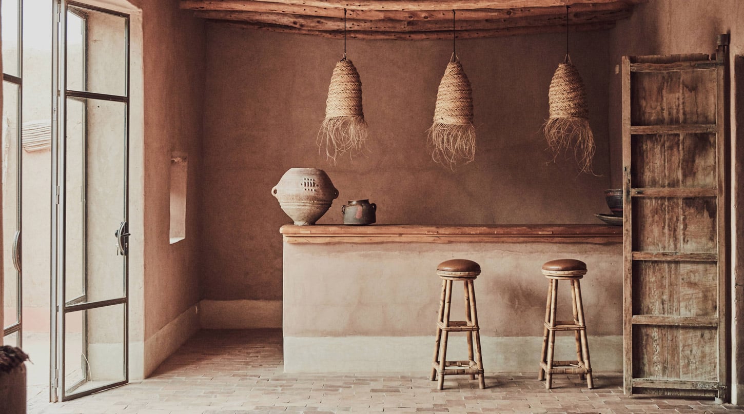 Berber Lodge, Africa — Earthy clay-walled bar interior with woven pendant lights, wooden bar stools, ceramic vessels, and rustic shelving in warm ochre tones.
