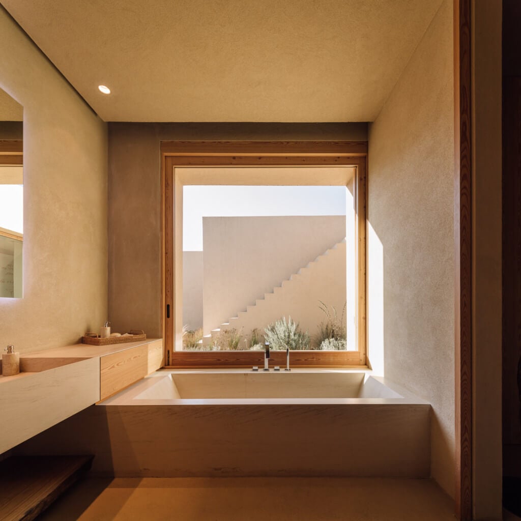 Melides Art, Portugal — Minimalist bathroom with concrete soaking tub, wooden cabinetry, and framed window overlooking staircase and desert landscape in warm natural light.