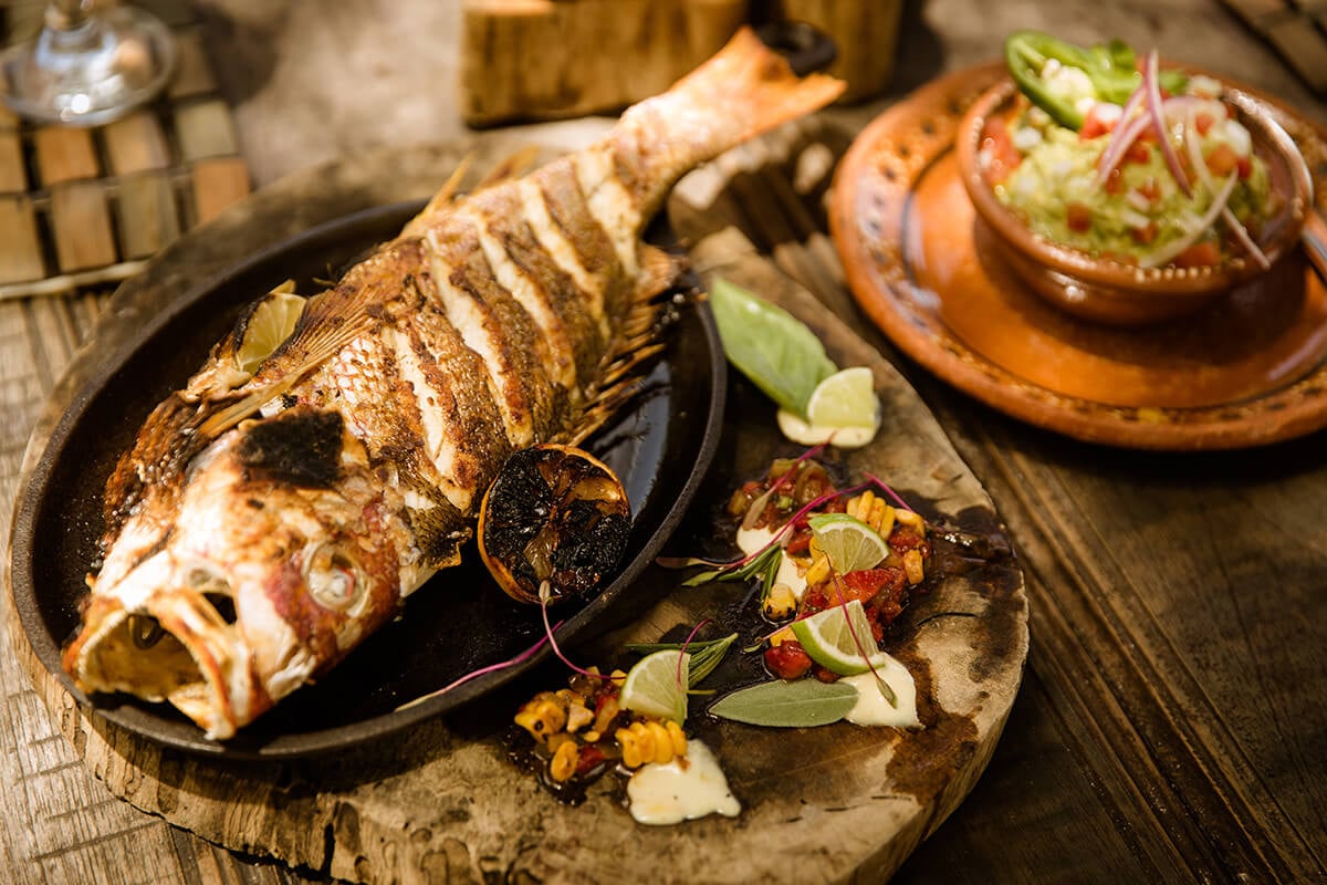 Be Tulum, Mexico — Grilled fish plated on black cast iron with lime, pickled vegetables, and edible flowers on wooden table.