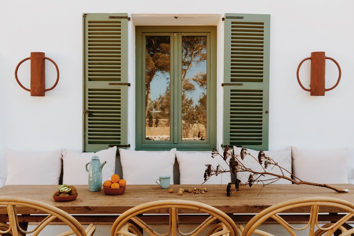 Menorca Experimental, Balearic Islands — Coastal dining setup with green louvered shutters, wooden table, white cushioned seating, terracotta accents, and dried botanicals overlooking Mediterranean landscape.