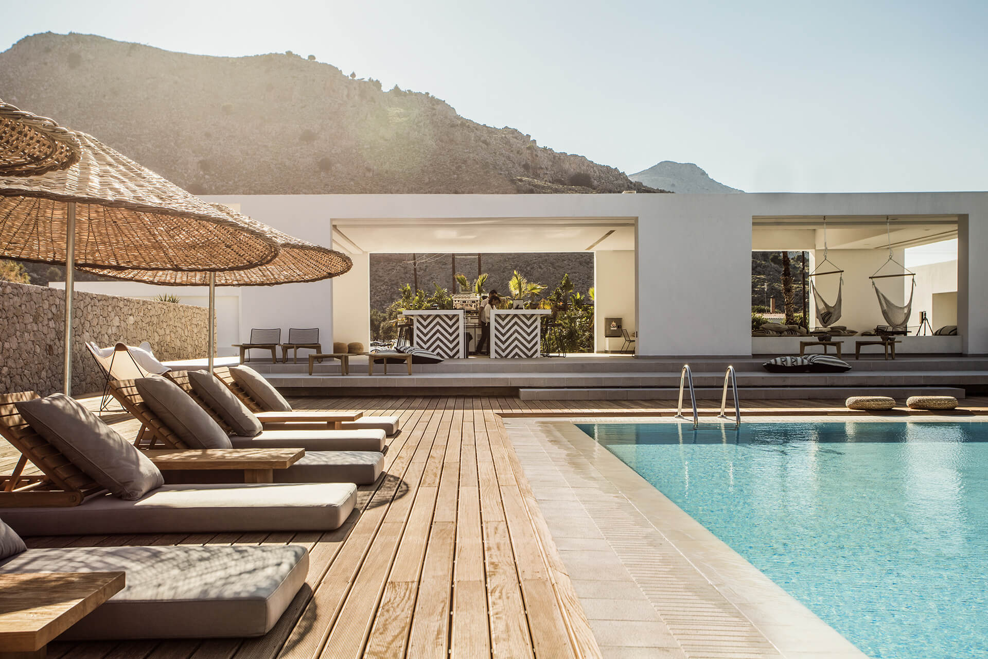 Casa Cook Rhodes, Rhodes, Greece — Modern poolside terrace with wooden decking, lounge chairs, woven umbrella, and minimalist white architecture overlooking Aegean mountains.