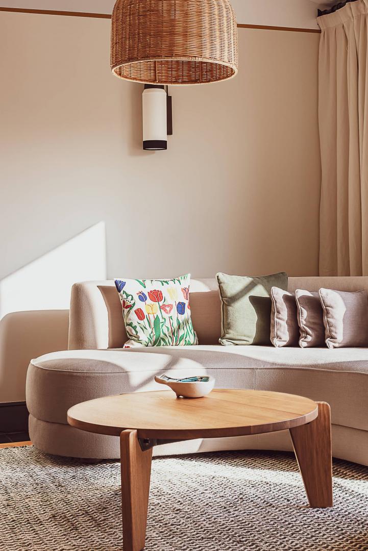 Hôtel Lou Pinet, Europe — Curved beige sofa with colorful floral pillow and woven rattan pendant lamp above wooden coffee table on natural fiber rug.