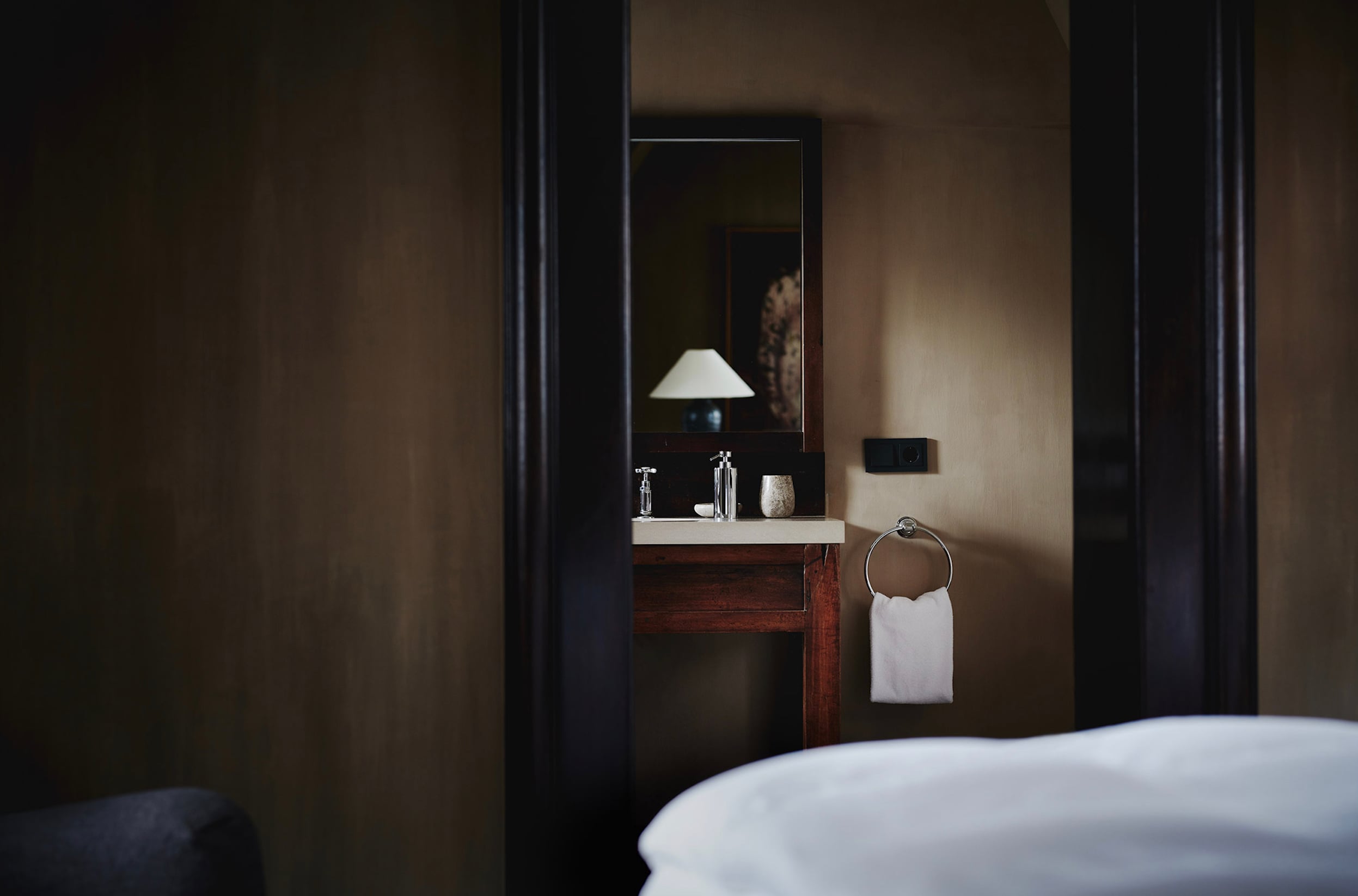Purs, Europe — Minimalist bathroom vanity with dark wood cabinetry, white basin, mirror, and pendant lamp beside bathtub in warm neutral tones.