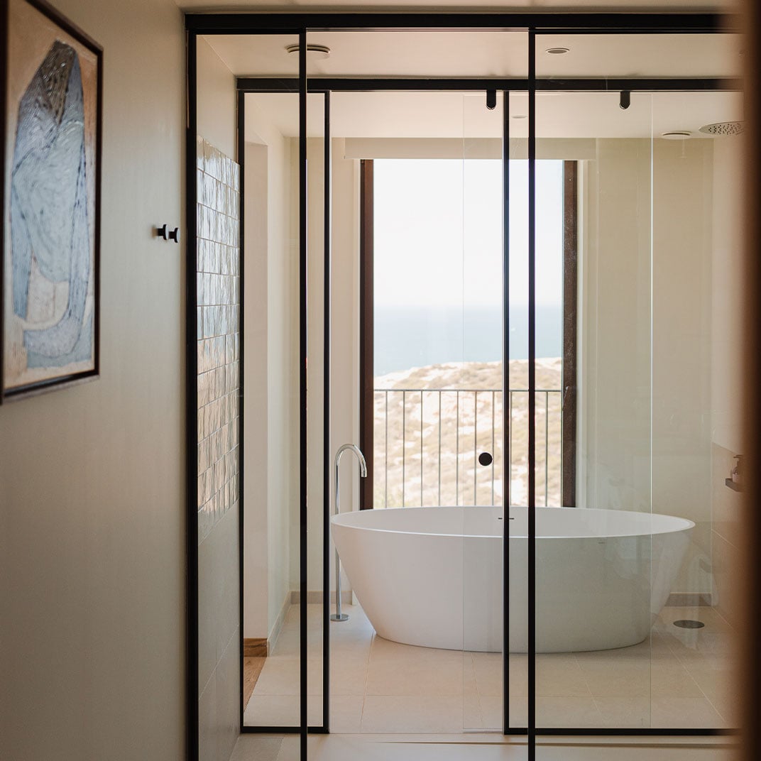 Aethos Ericeira, Portugal — Modern bathroom with freestanding oval tub, black-framed glass enclosure, coastal landscape view through floor-to-ceiling windows.